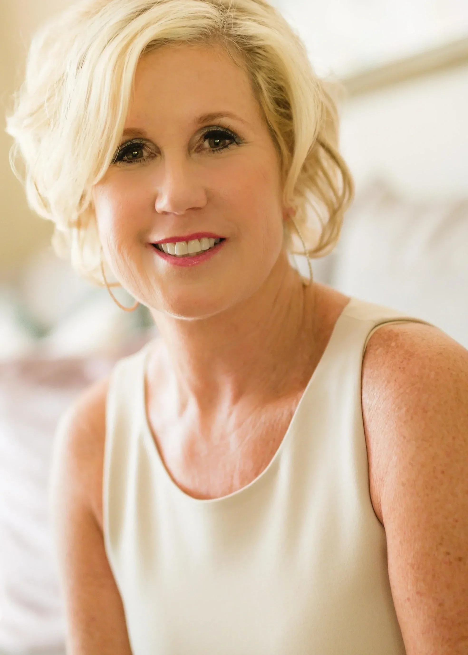 A woman with blonde hair smiling, wearing a sleeveless cream-colored top, in a bright indoor setting.