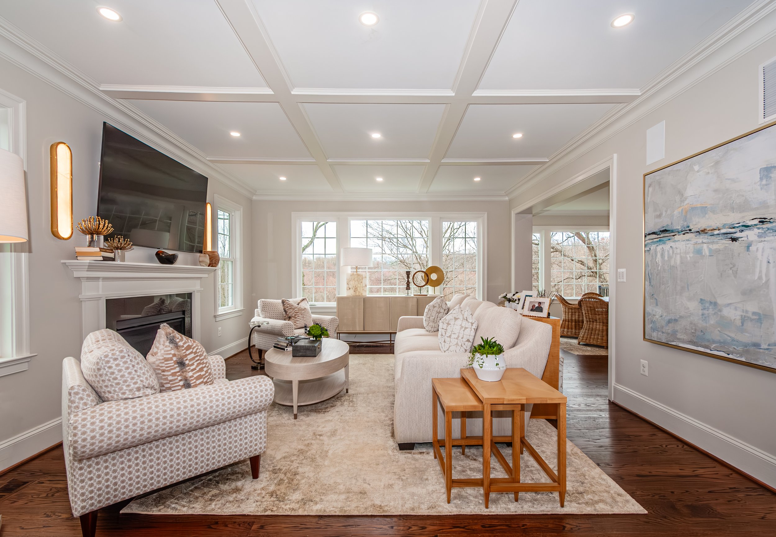 Living room with white walls, a television above a fireplace, beige sofas, armchair, and wooden and decorated accents, large windows at the back.