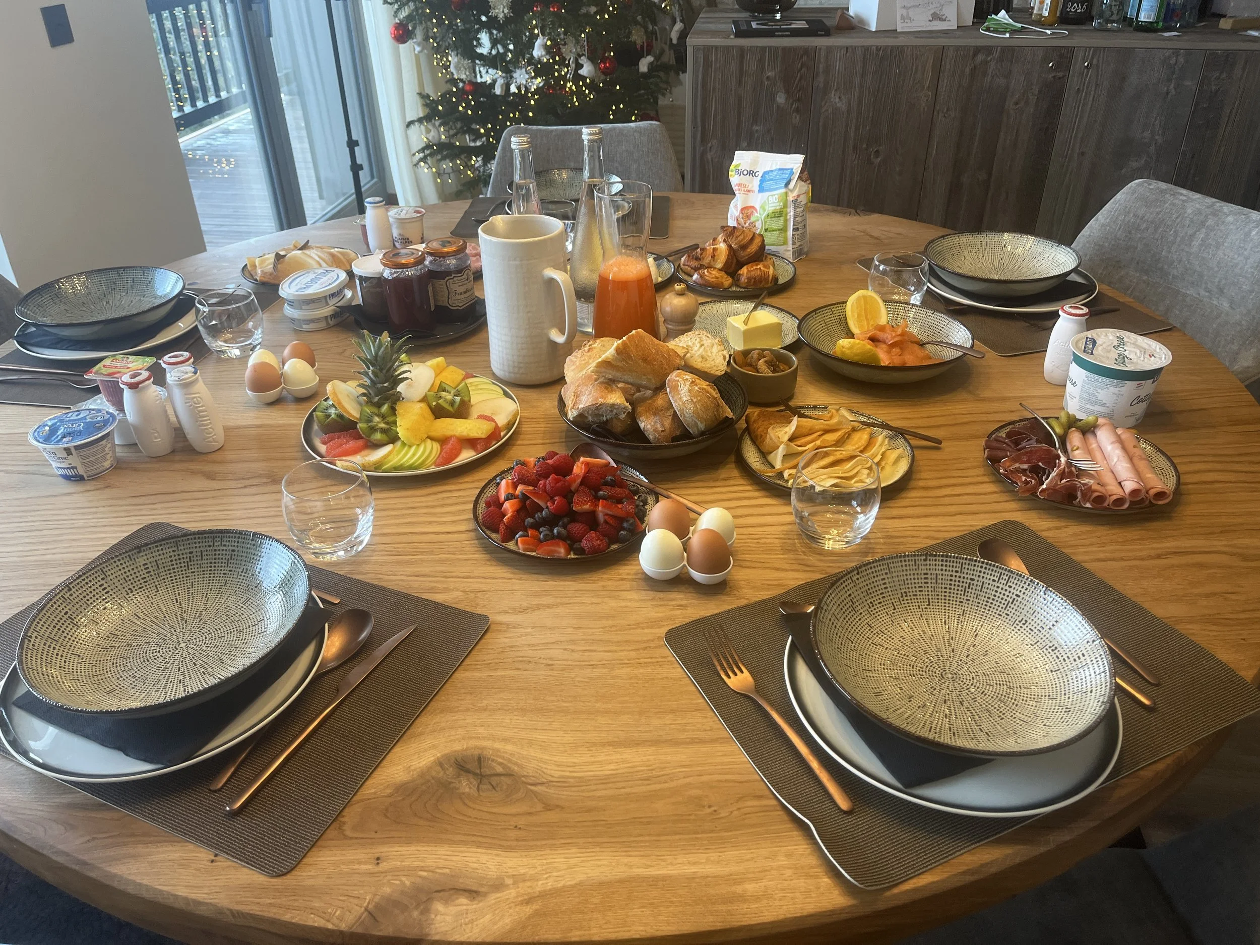 Table de petit-déjeuner avec fruits, œufs, viennoiseries, charcuterie, yaourt, jus d'orange, et autres aliments, décorée pour Noël avec un sapin en arrière-plan.