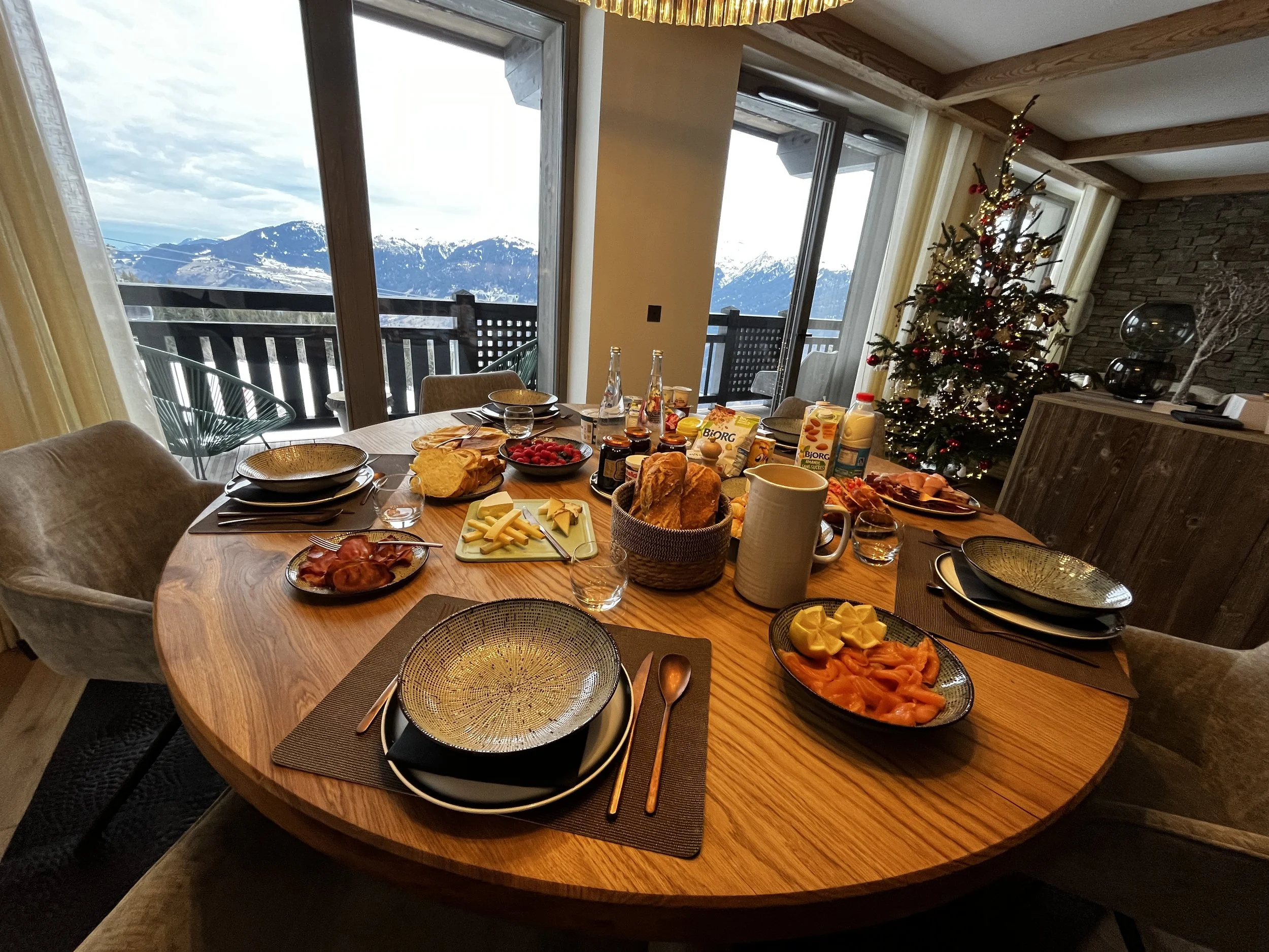 Table de petit déjeuner avec divers plats, fromages, fruits, pain, et boissons, dans une salle avec vue sur une montagne enneigée et un sapin de Noël décoré.