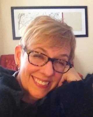 A smiling woman with short blonde hair and glasses, sitting on a red chair with a decorated wall in the background.