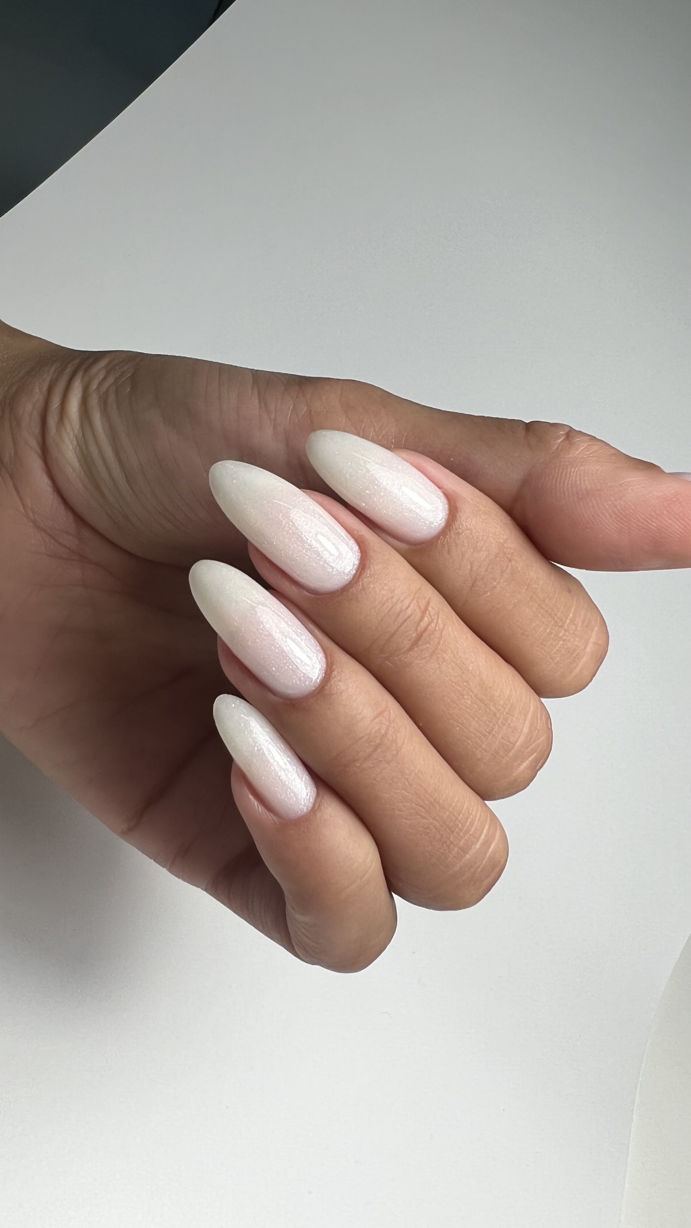 Close-up of a hand with neatly manicured nails painted pale pink with a glittery finish, resting on a white surface.