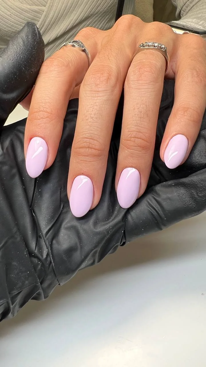 Close-up of a hand with pastel pink oval-shaped manicured nails, two silver rings on the middle and ring fingers, and the hand resting on a black glove.