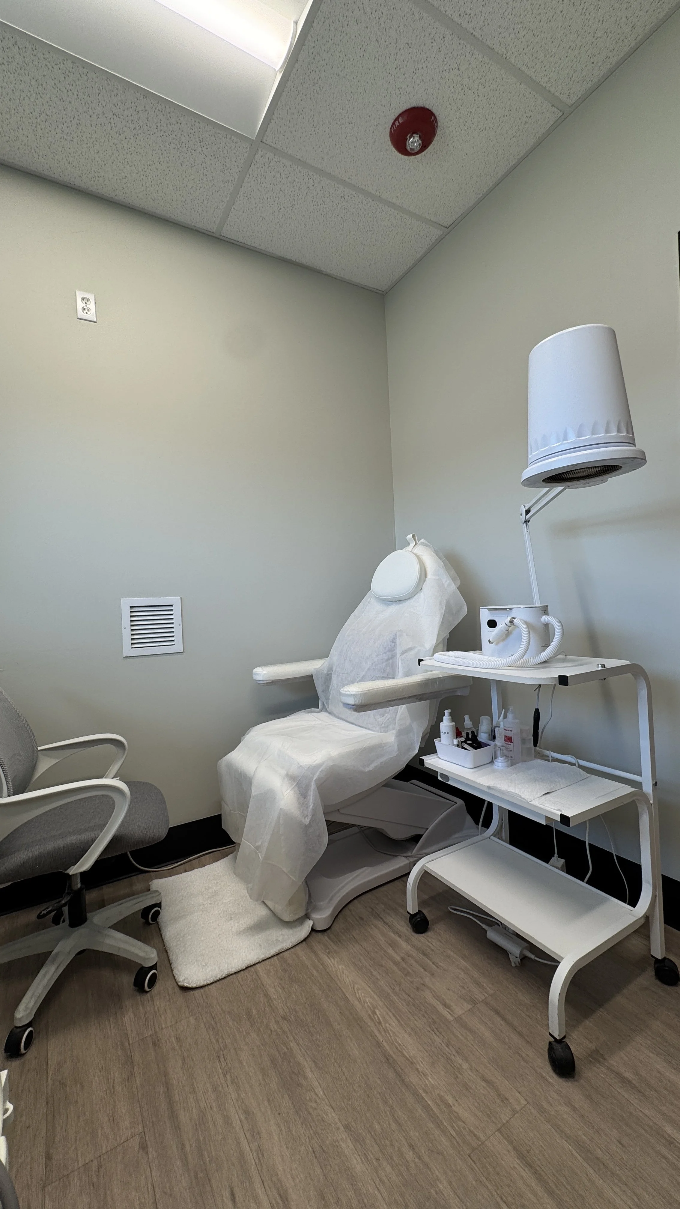 Medical examination room with a white reclining chair covered with paper, a white rolling cart with supplies, and a lamp.