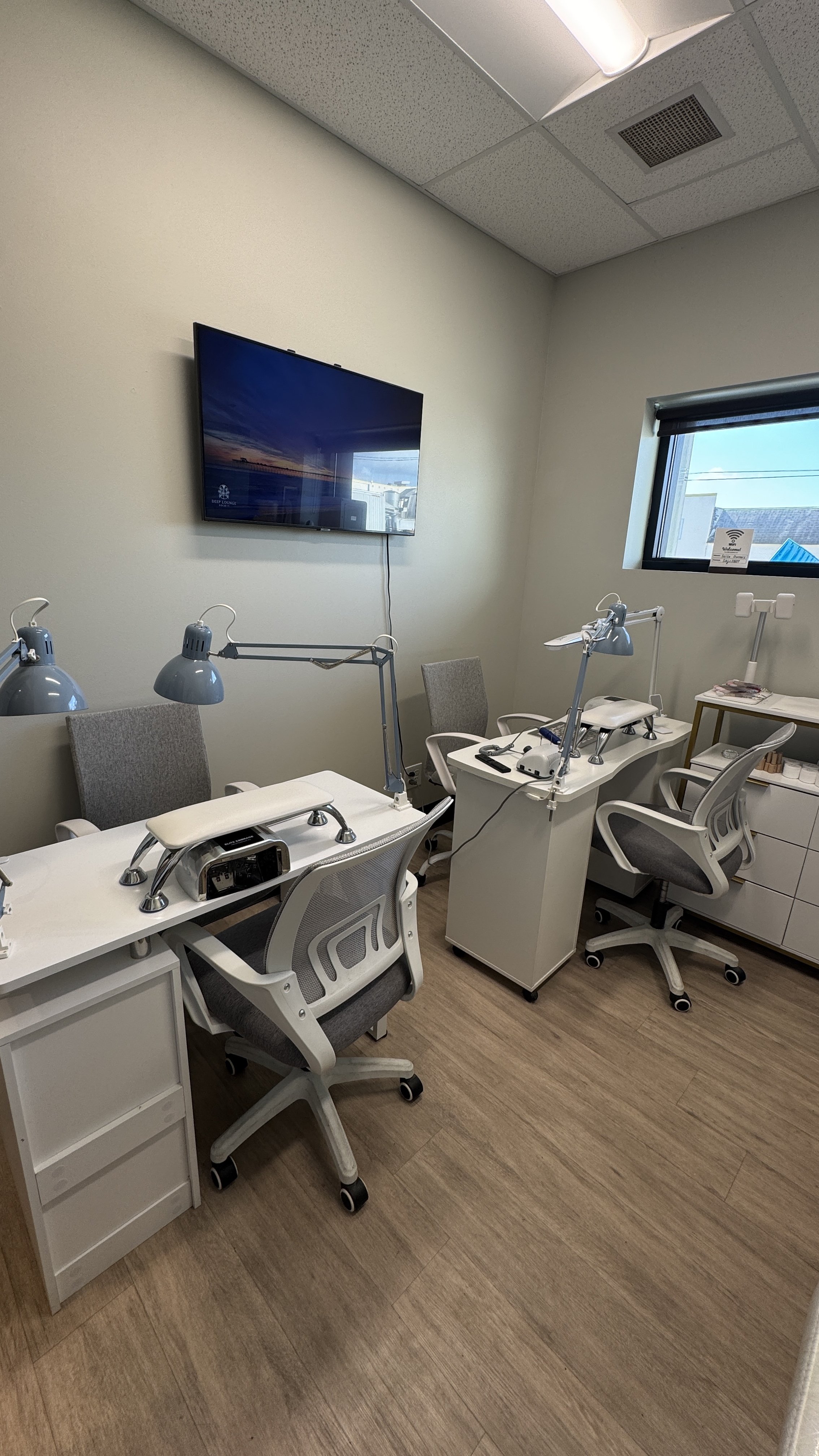A room with four desks, each equipped with a lamp, a magnifying light, and ergonomic chairs. There is a wall-mounted TV, a window with blinds, and a cabinet with supplies. The floor is wood, and the ceiling has a fluorescent light and an air vent.