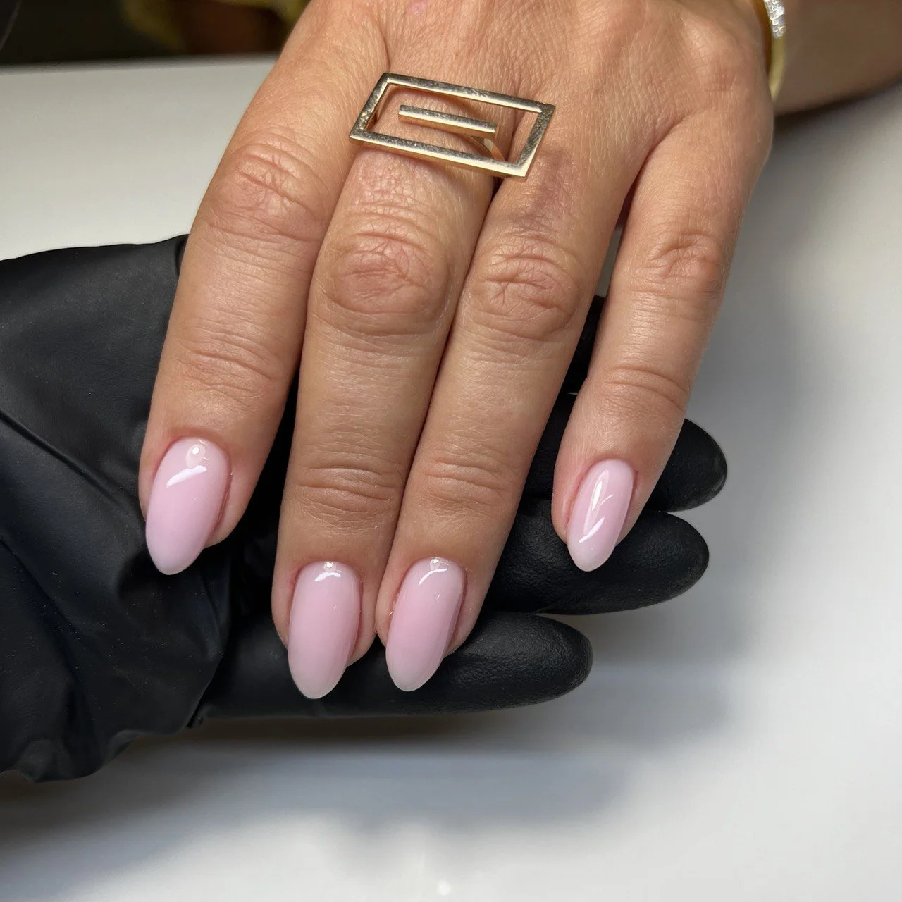 Close-up of a hand with pink ombre nails, wearing a large gold ring with a geometric design, held over a white surface. The person is wearing a black glove on the hand with the nails.