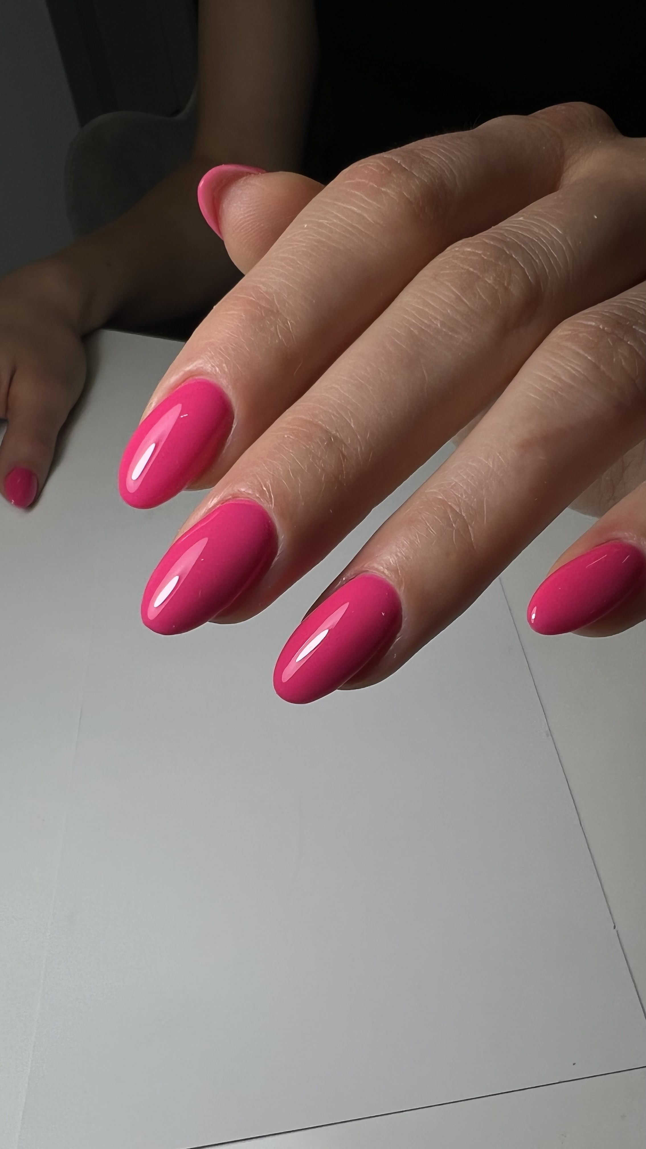 Close-up of a hand with pink manicured nails, resting on a white surface, with a person in the background.