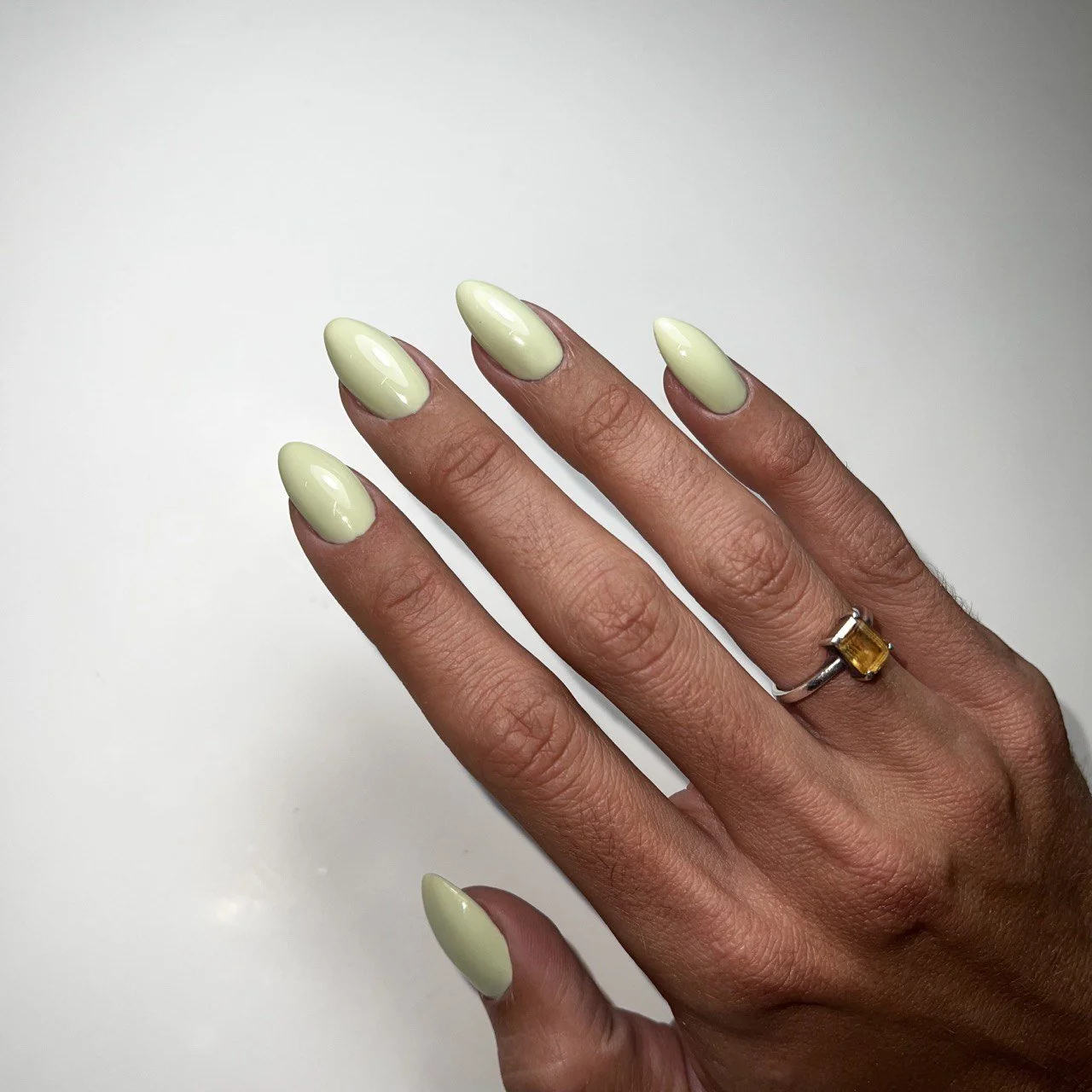 A hand with well-manicured nails painted in a light green color, wearing a ring with a yellow gemstone on the ring finger, against a plain white background.