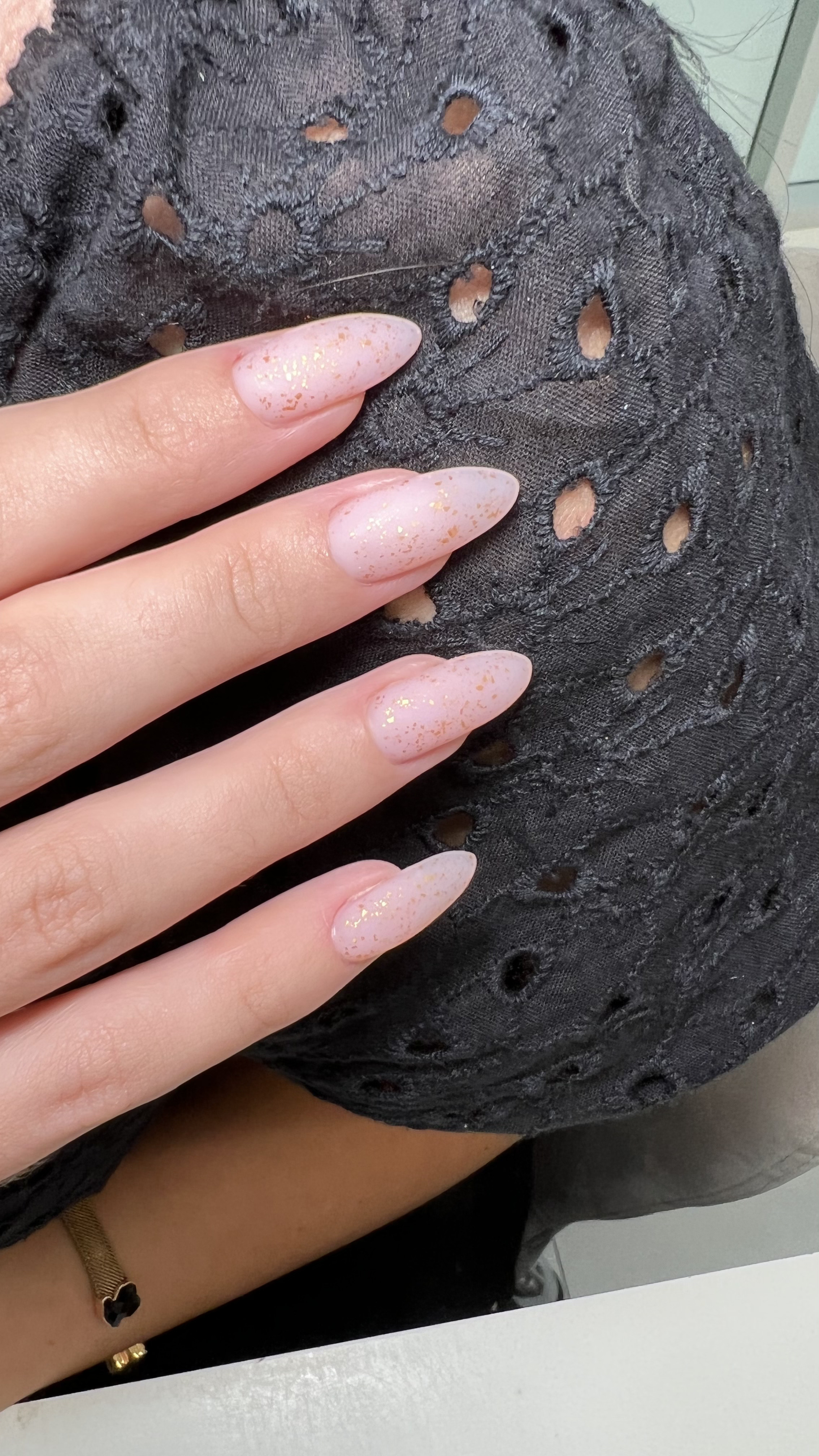 Close-up of a hand with long, pointed nails painted in a light pink color with gold speckles, resting on a distressed black fabric with circular cutouts.