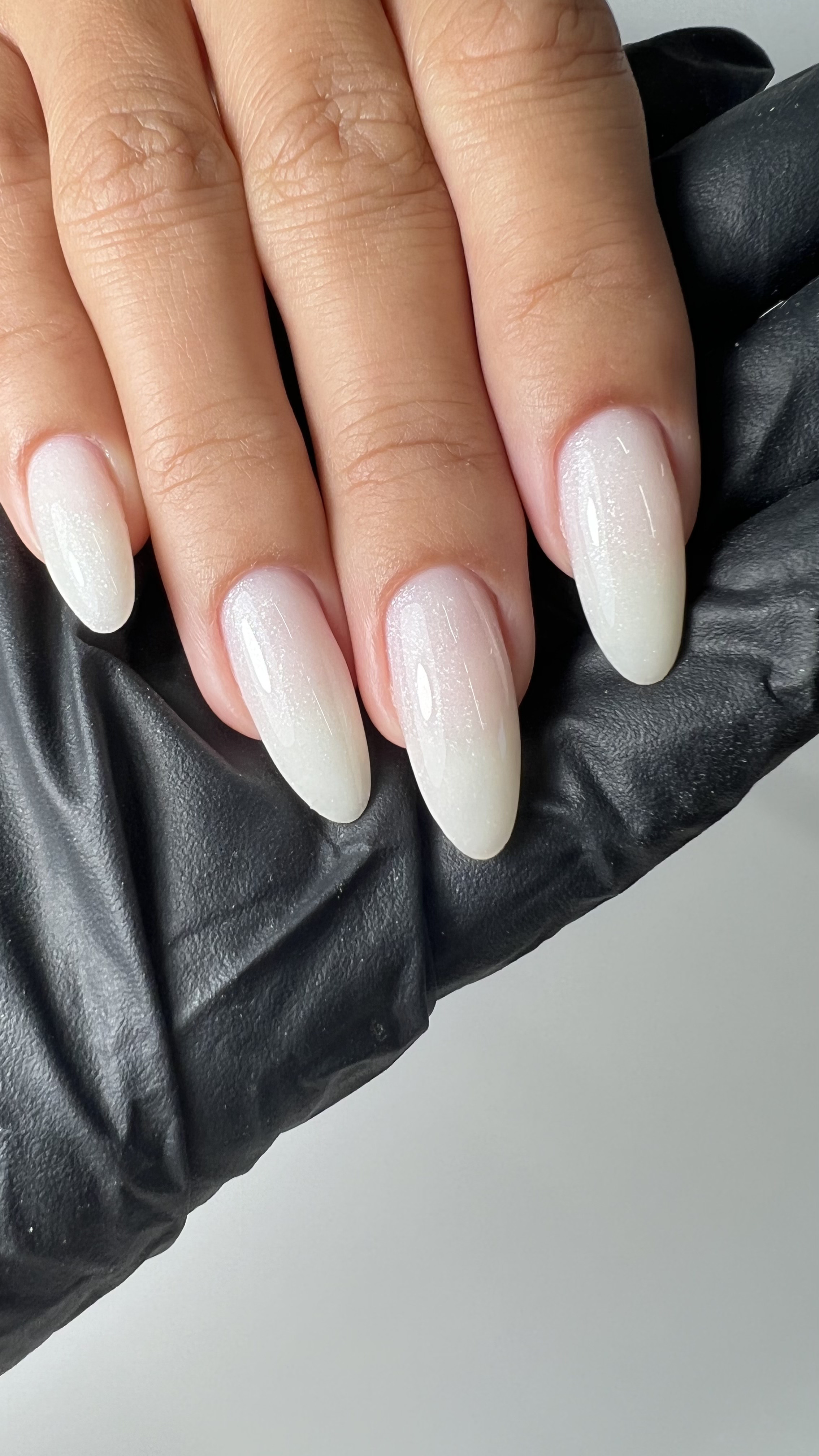 Close-up of a hand with long, almond-shaped acrylic nails painted in a pearly white with a subtle shimmer, resting on a black leather surface.