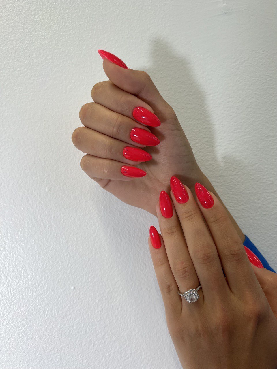 Close-up of a hand with bright red, elongated, glossy nails, and a diamond engagement ring on the ring finger, against a plain white background.