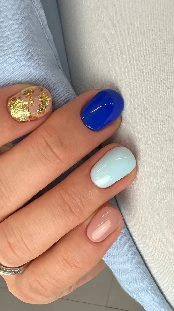 Close-up of a person's hand with painted nails in gold, blue, white, and nude colors, resting on light-colored fabric.