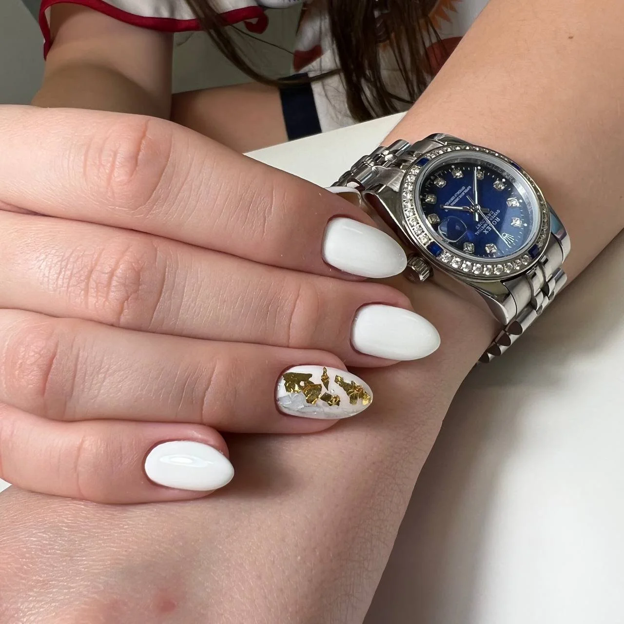 Close-up of a woman's hand showing a large silver wristwatch with a blue face and diamond accents, resting on another person's arm. The person's nails are painted white, with one nail decorated with gold foil patches.