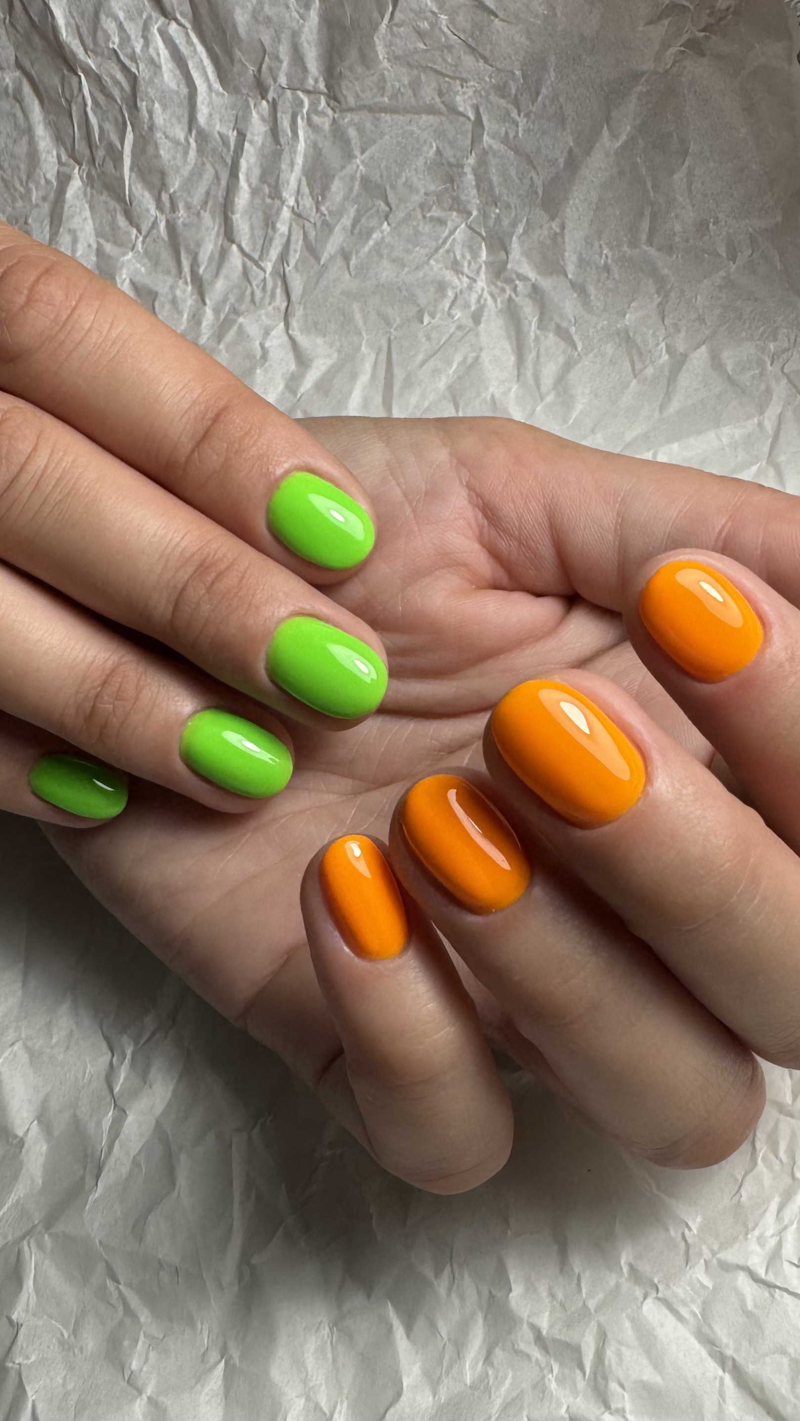 Close-up of hands with vibrant green and orange manicured nails, resting on crumpled white paper.