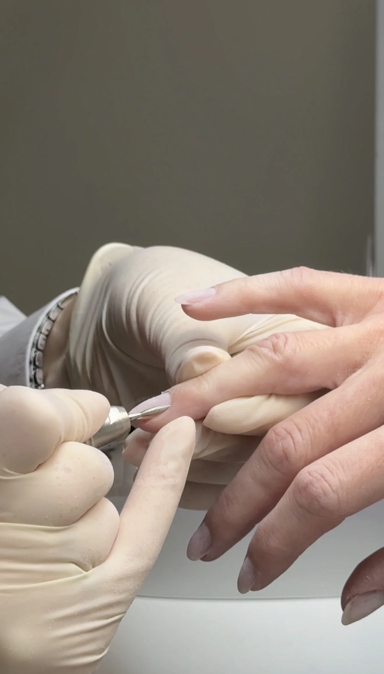 A nail technician is applying a clear coat to a client's fingernails with a nail drill, wearing gloves.
