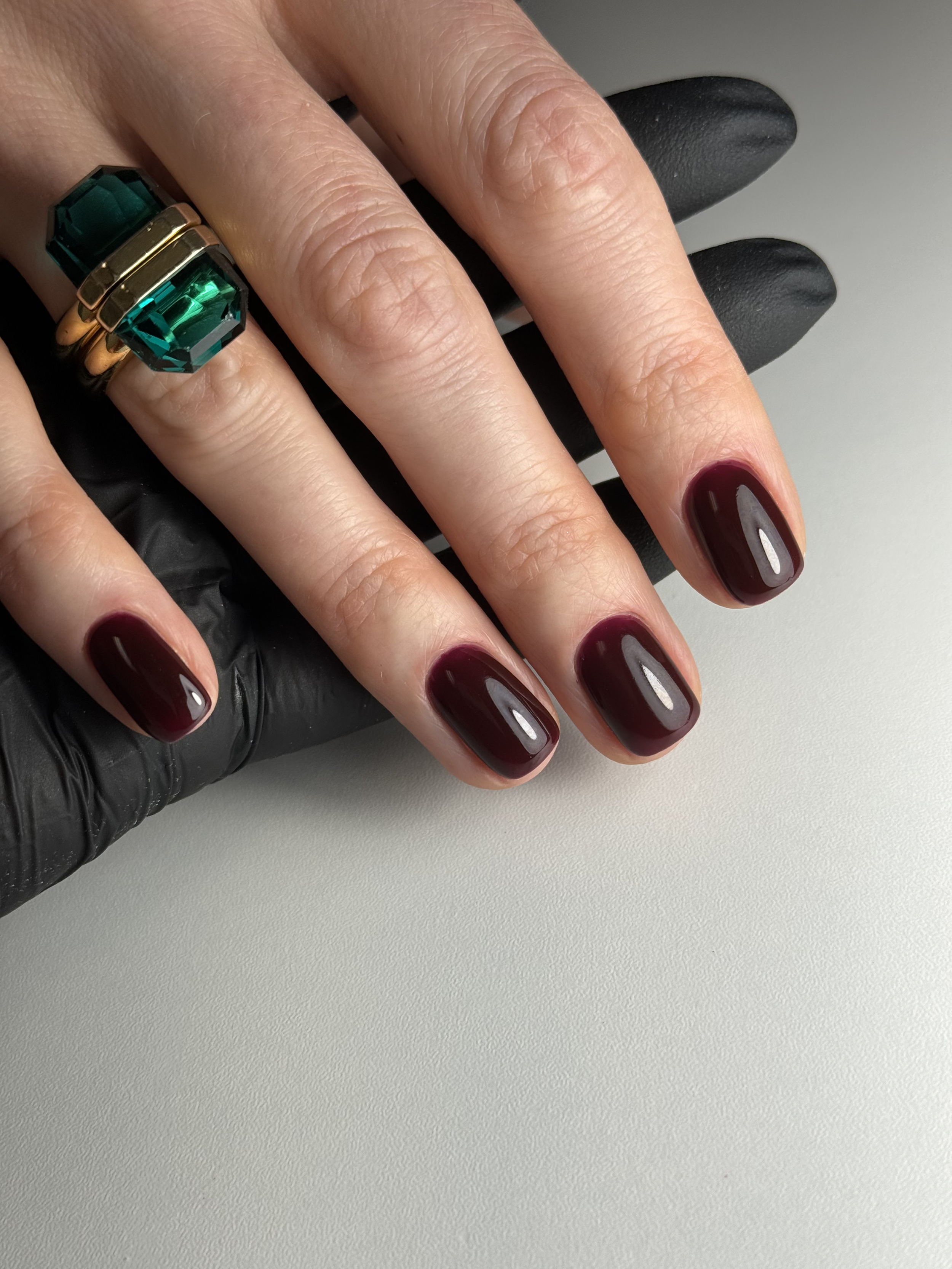 Close-up of a hand with dark red manicured nails, wearing a large green gemstone ring on the middle finger and a black glove on the index finger.