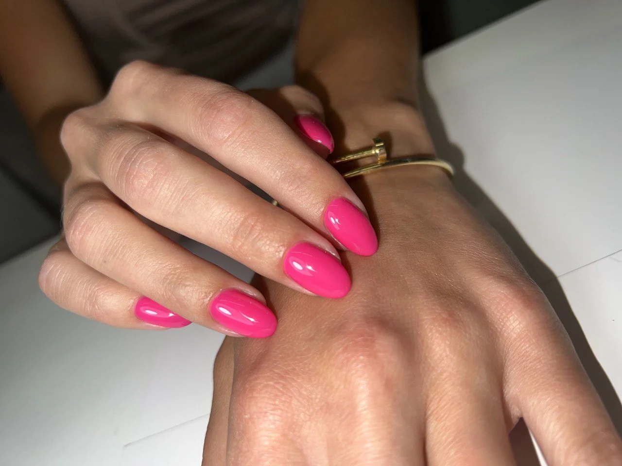 Close-up of a woman's hand with pink manicured nails, wearing a gold bracelet and a gold ring, resting on another hand.