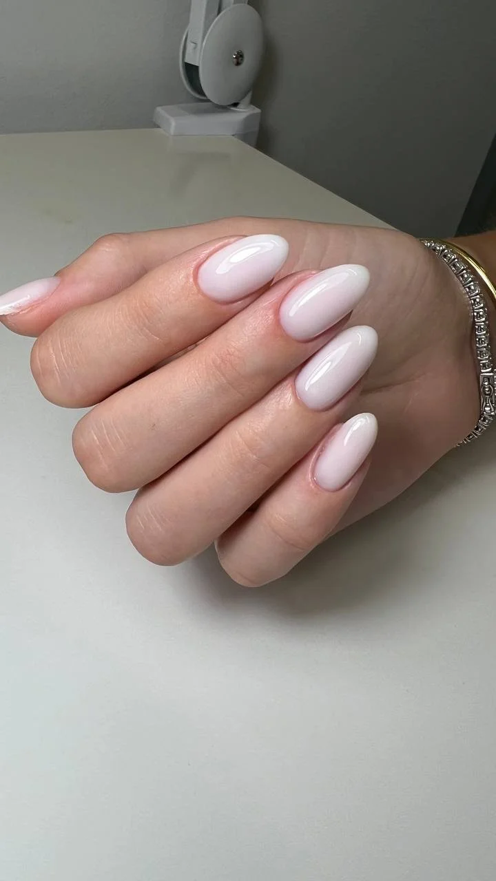 Close-up of a hand with neatly manicured, oval-shaped nails painted in a light pink or nude color, resting on a white surface.