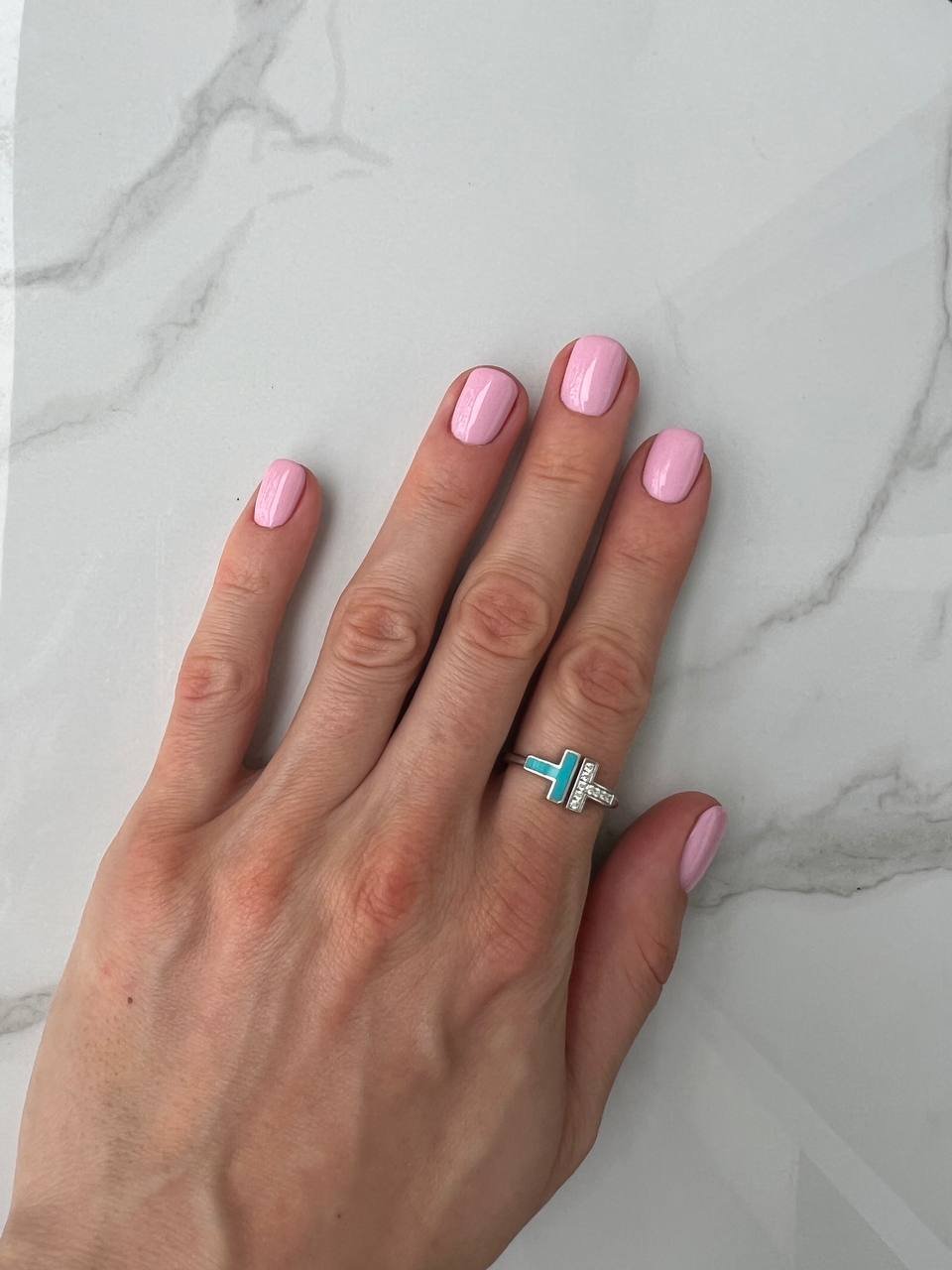 A hand with pink nail polish wearing a ring with a letter 'T' on it, resting on a white marble surface.