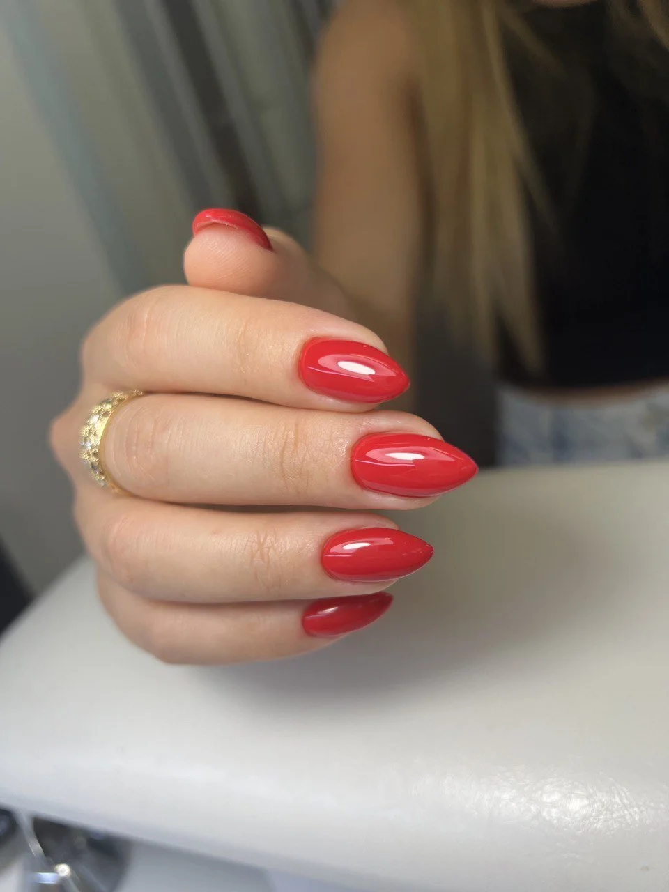 Close-up of a woman's hand with bright red, almond-shaped manicured nails, wearing a gold ring.