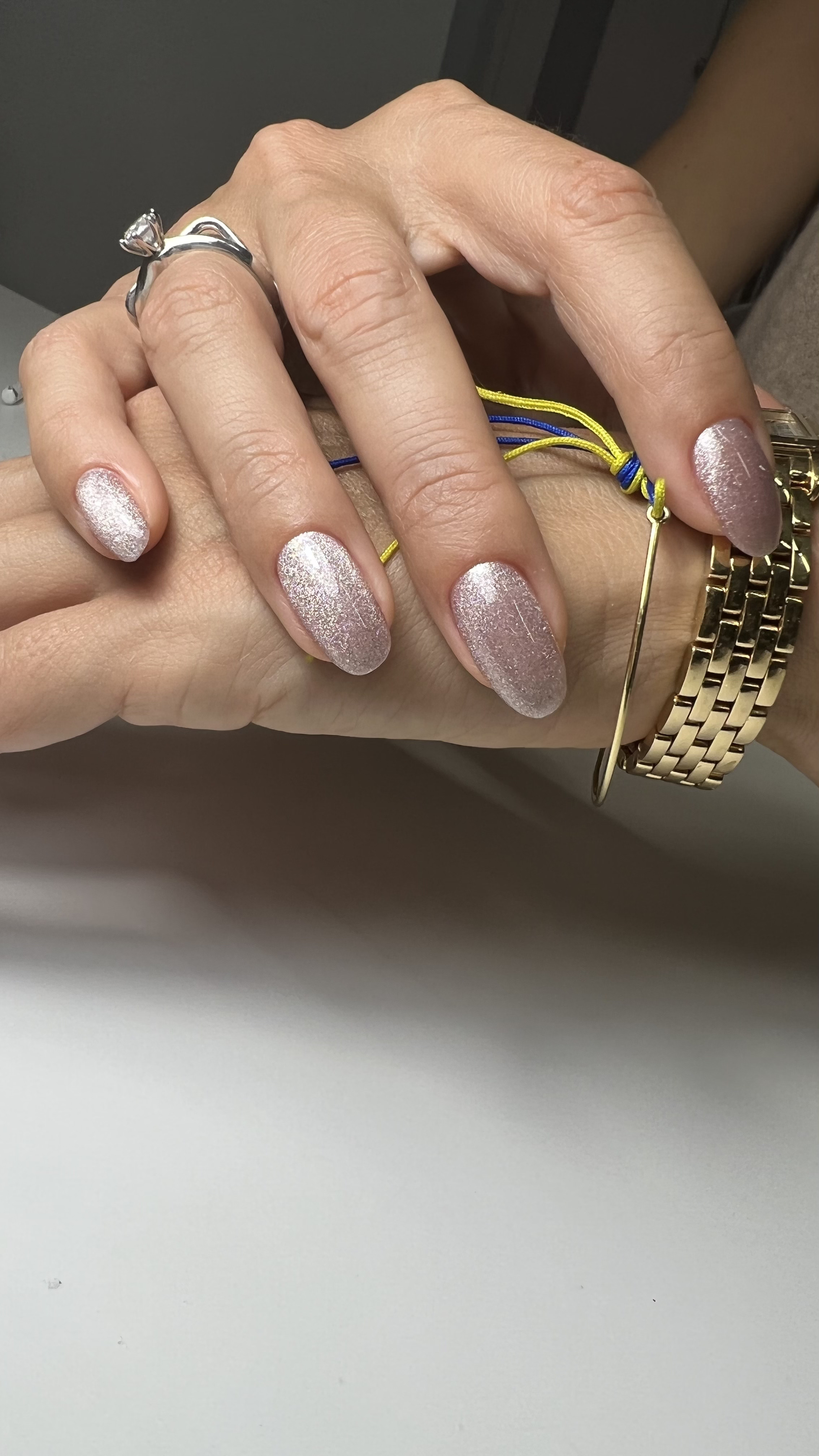 Close-up of a person's hand with silver glitter nail polish, wearing a silver ring, a gold watch, and a bracelet. The hand is holding a yellow and blue string bracelet.