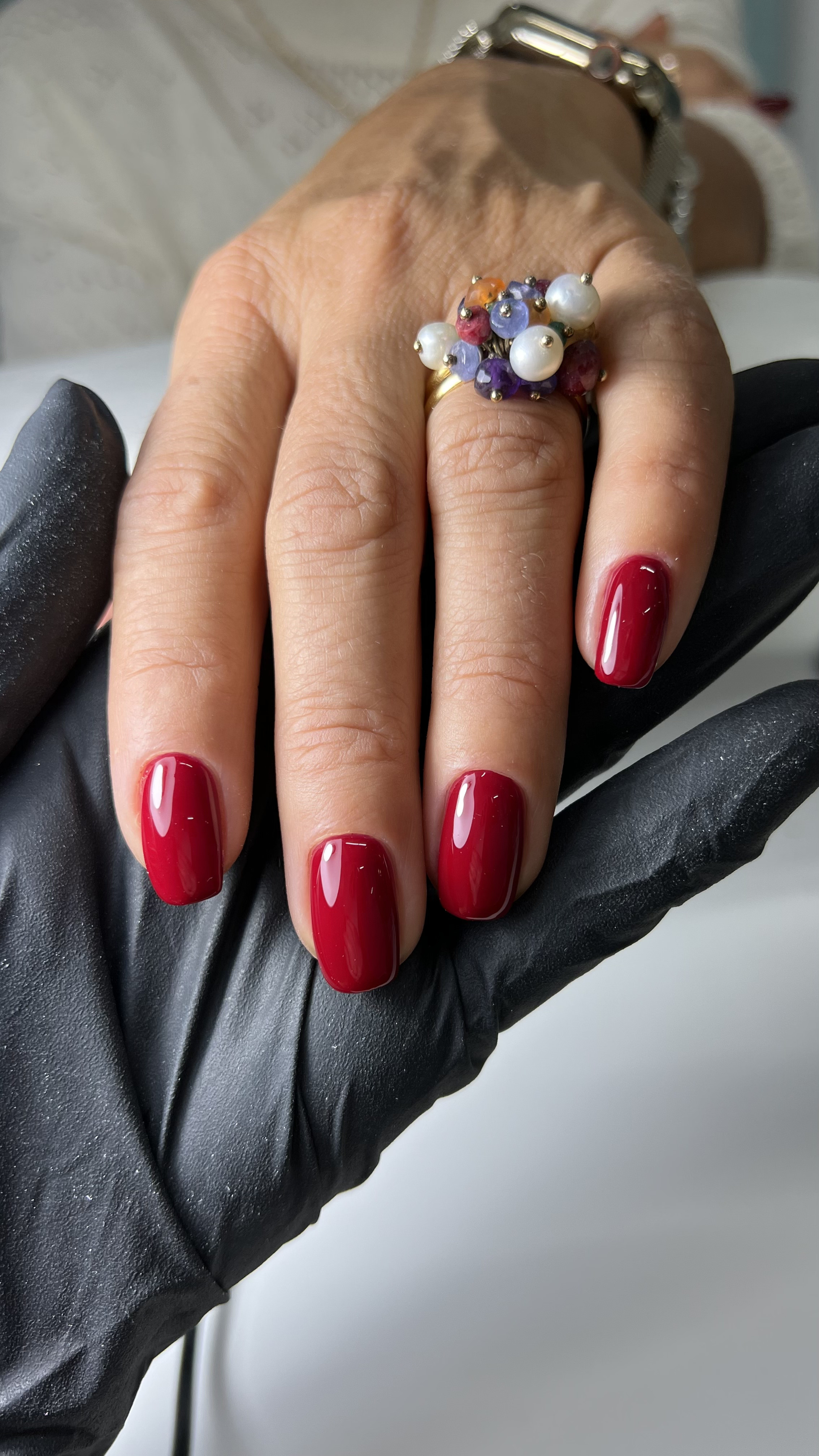 Close-up of a hand with five fingers, showing red nail polish and a gold ring with colorful pearls and small gemstones. A silver watch is visible on the wrist. The hand is held against a black gloved hand, against a neutral background.