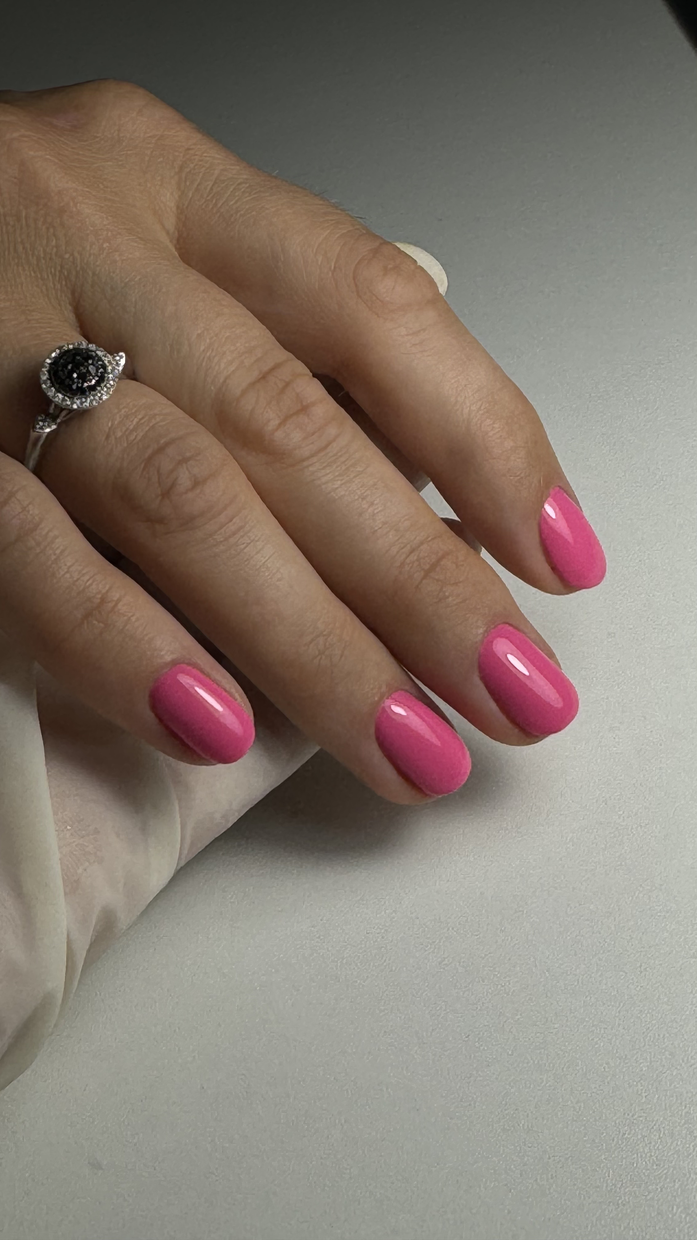 Close-up of a hand with pink manicured nails, wearing a ring with a dark gemstone surrounded by smaller clear stones.