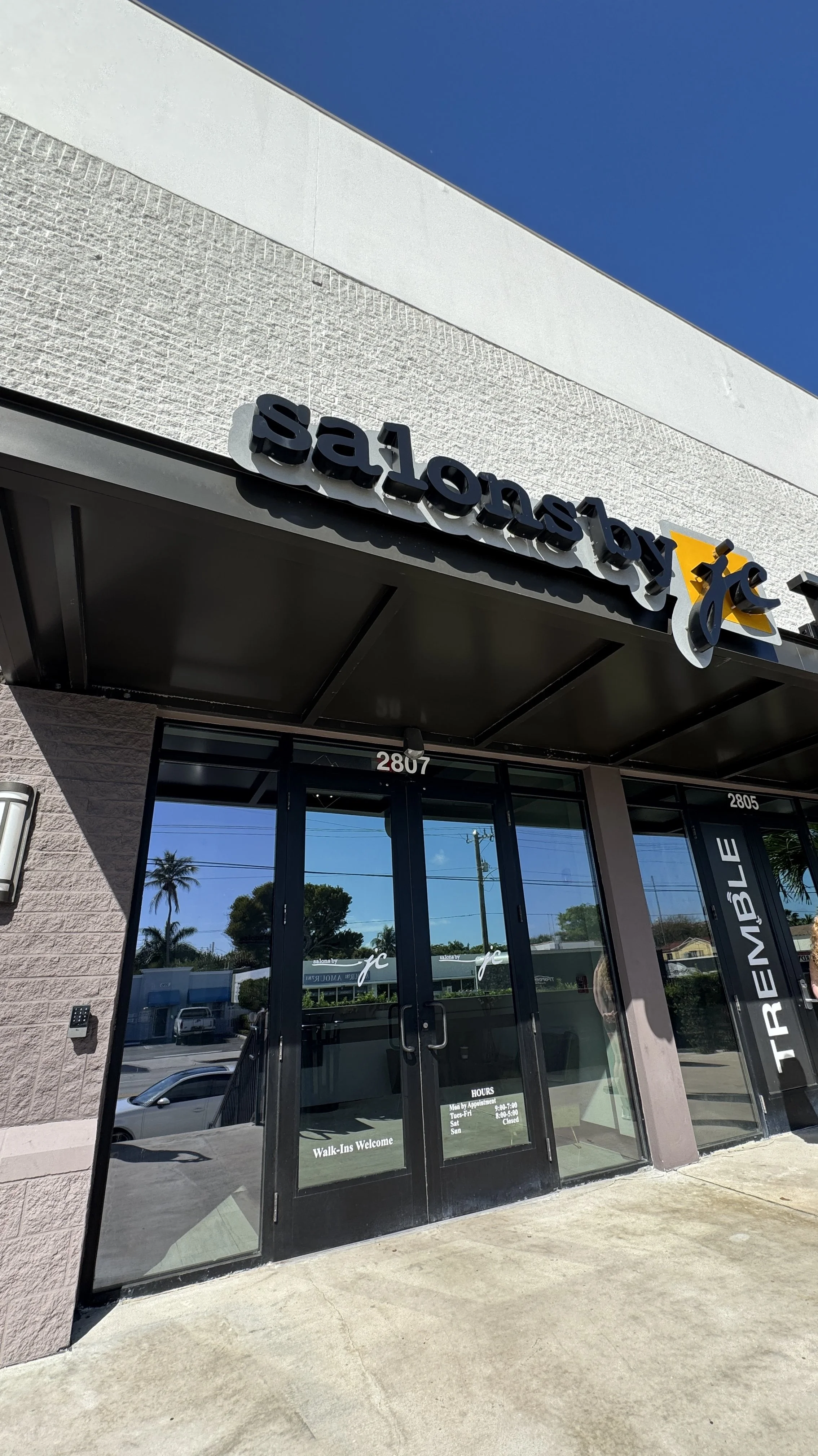 Exterior of a salon called 'Salonby jc' with glass doors and black framing, reflecting parked cars and palm trees outside.