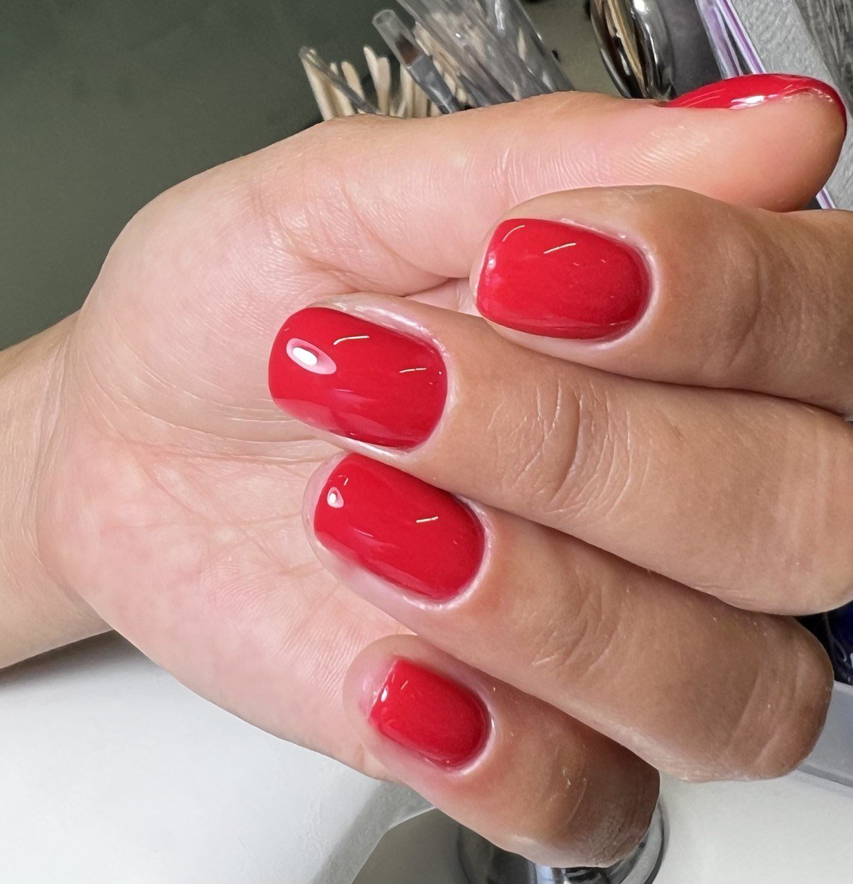 Close-up of a person's hand showing freshly manicured nails painted in bright red polish, with a background of nail tools.