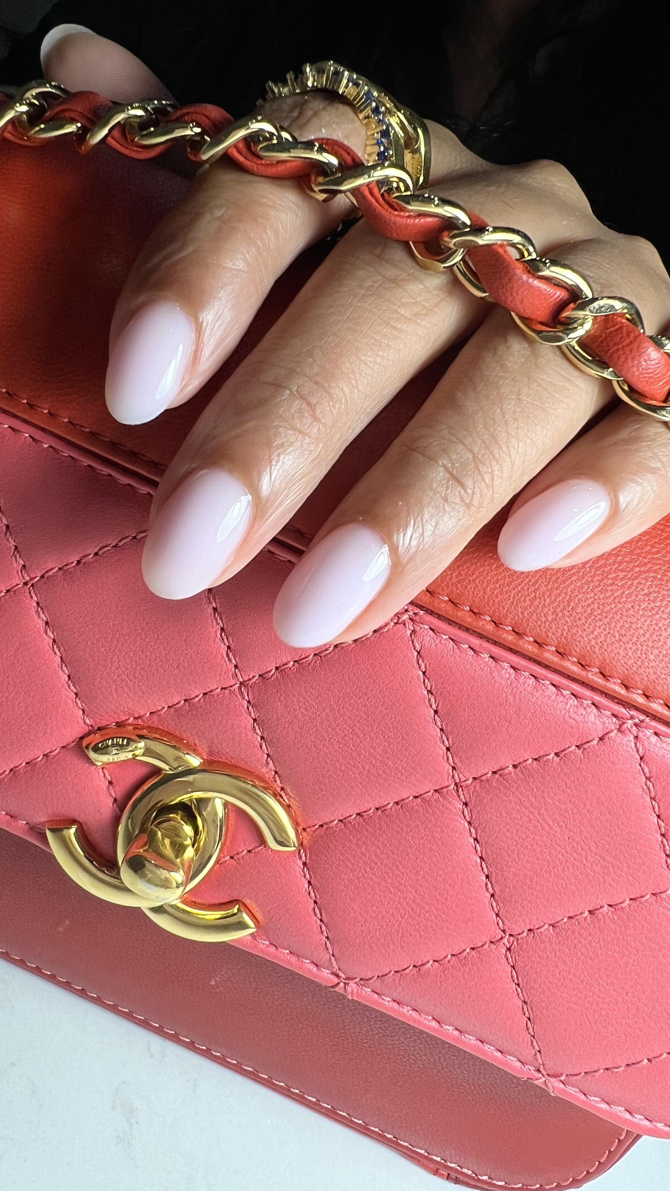 Close-up of a pink quilted handbag with a gold chain strap and a gold logo clasp. A hand with well-manicured, oval-shaped, white-painted nails rests on the bag, wearing a gold bracelet with dark blue gemstones.