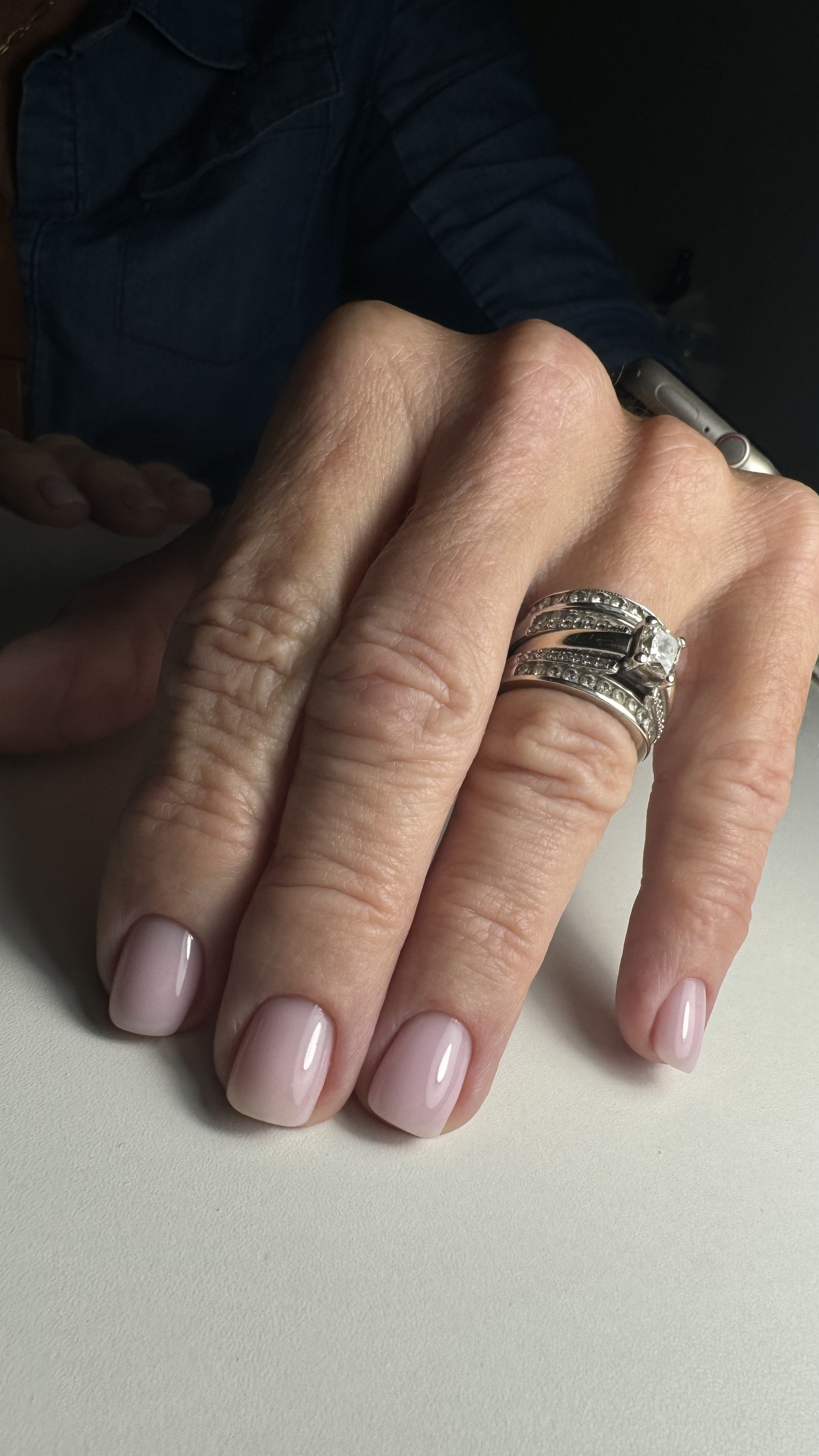 Close-up of a woman's hand with painted nails, showing multiple silver rings with gemstones and a large diamond. Part of a person's arm in a blue denim shirt is visible in the background.