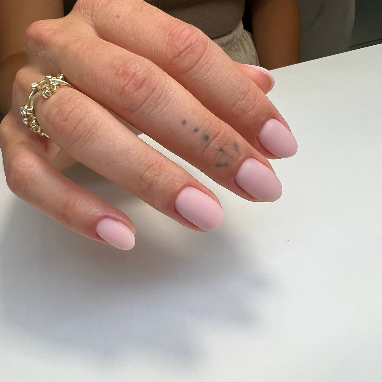 Close-up of a woman's left hand with pink nail polish, gold rings, and a small tattoo on the finger.