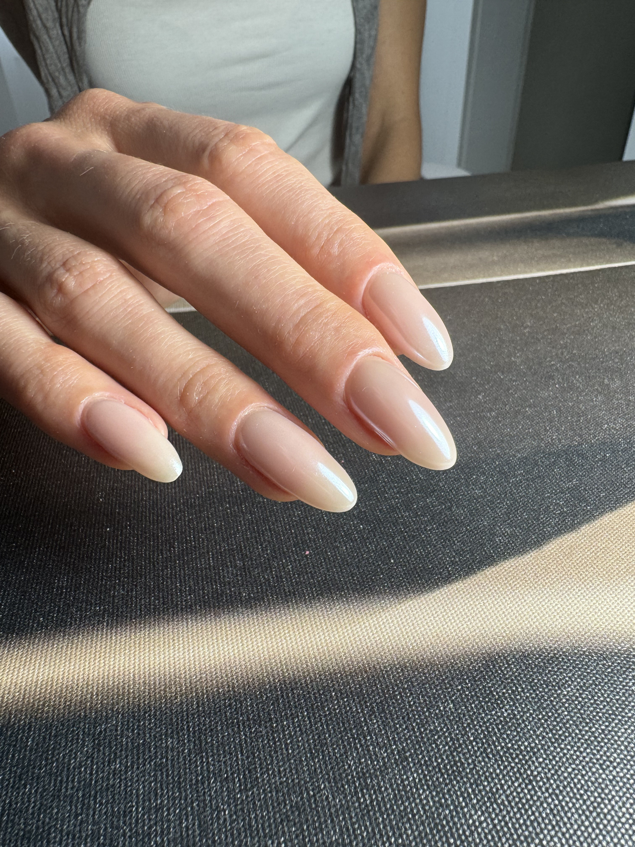 Close-up of a woman's hand with natural long, almond-shaped, glossy, nude-colored nails resting on a black surface.