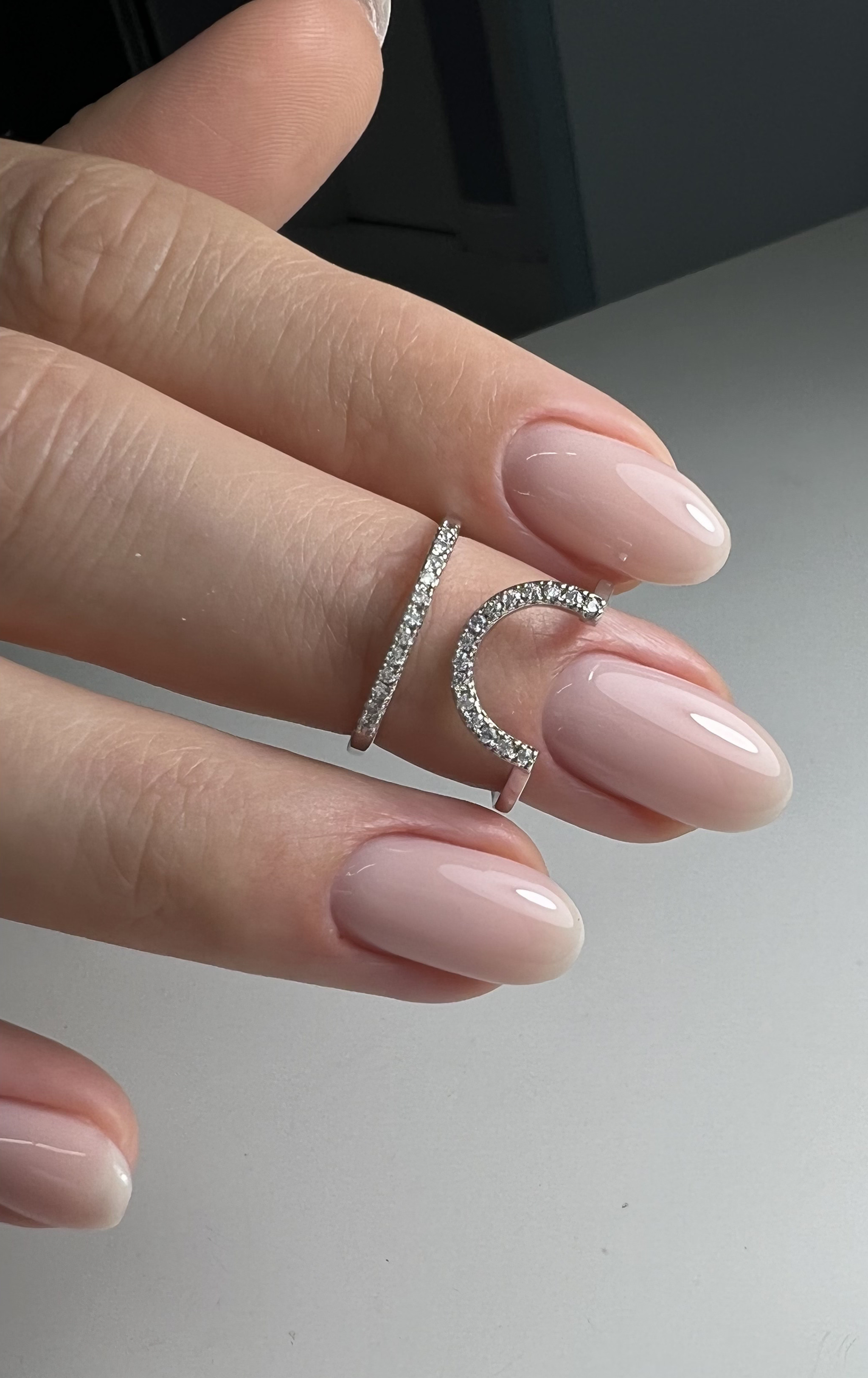 Close-up of a hand with light pink nails and two silver rings, one with a straight band and the other with a curved open design, both encrusted with small rhinestones.