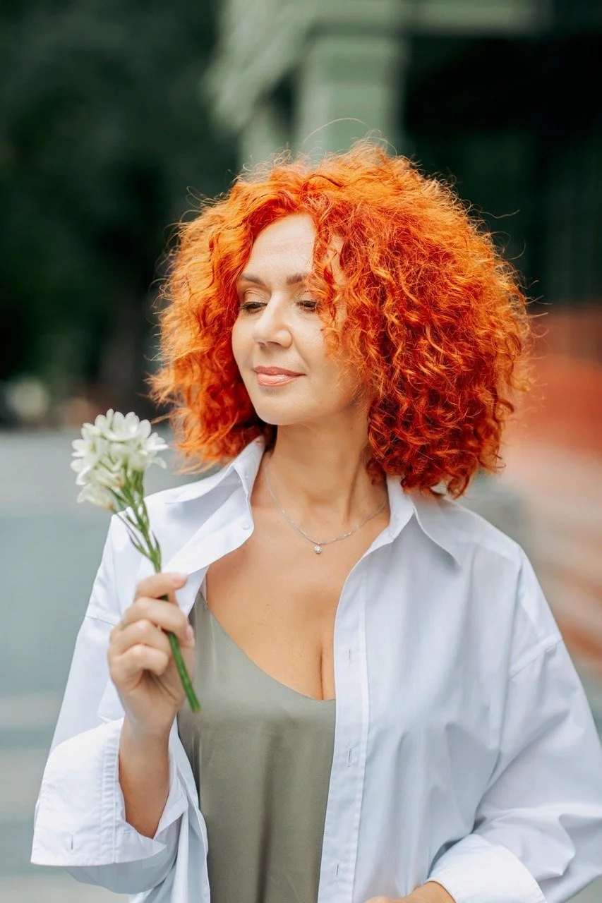 A woman with bright curly red hair, wearing a white shirt and a necklace, holding a small bouquet of white flowers and looking at them thoughtfully outdoors.