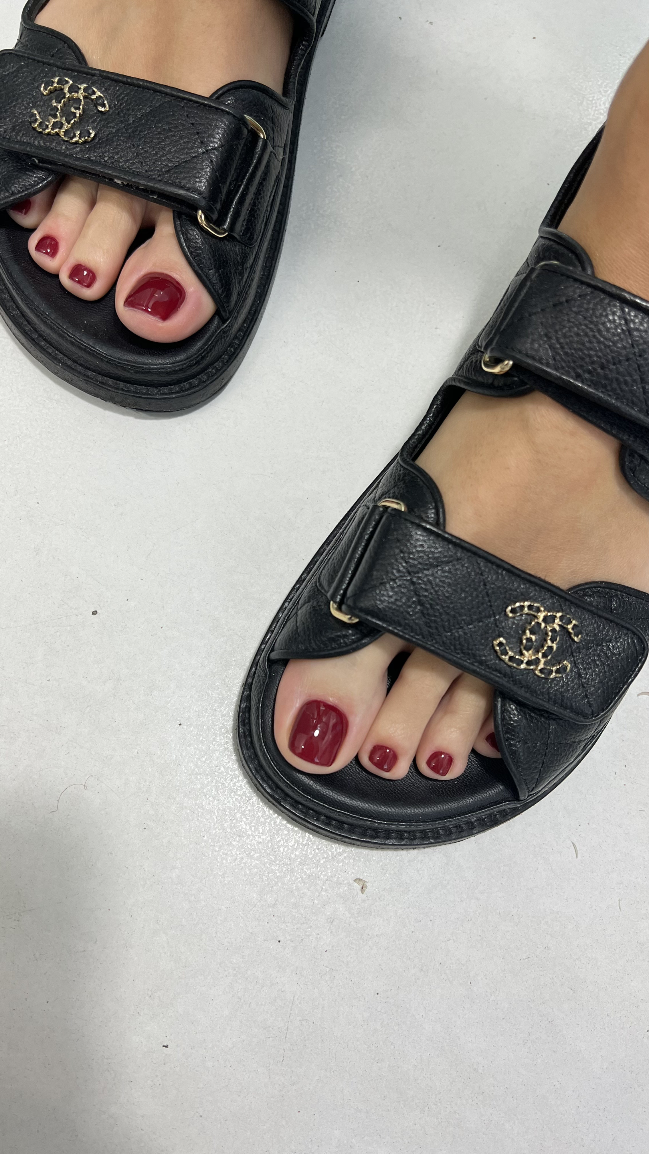 pair of feet wearing black sandals with a gold logo and red nail polish, standing on a white surface