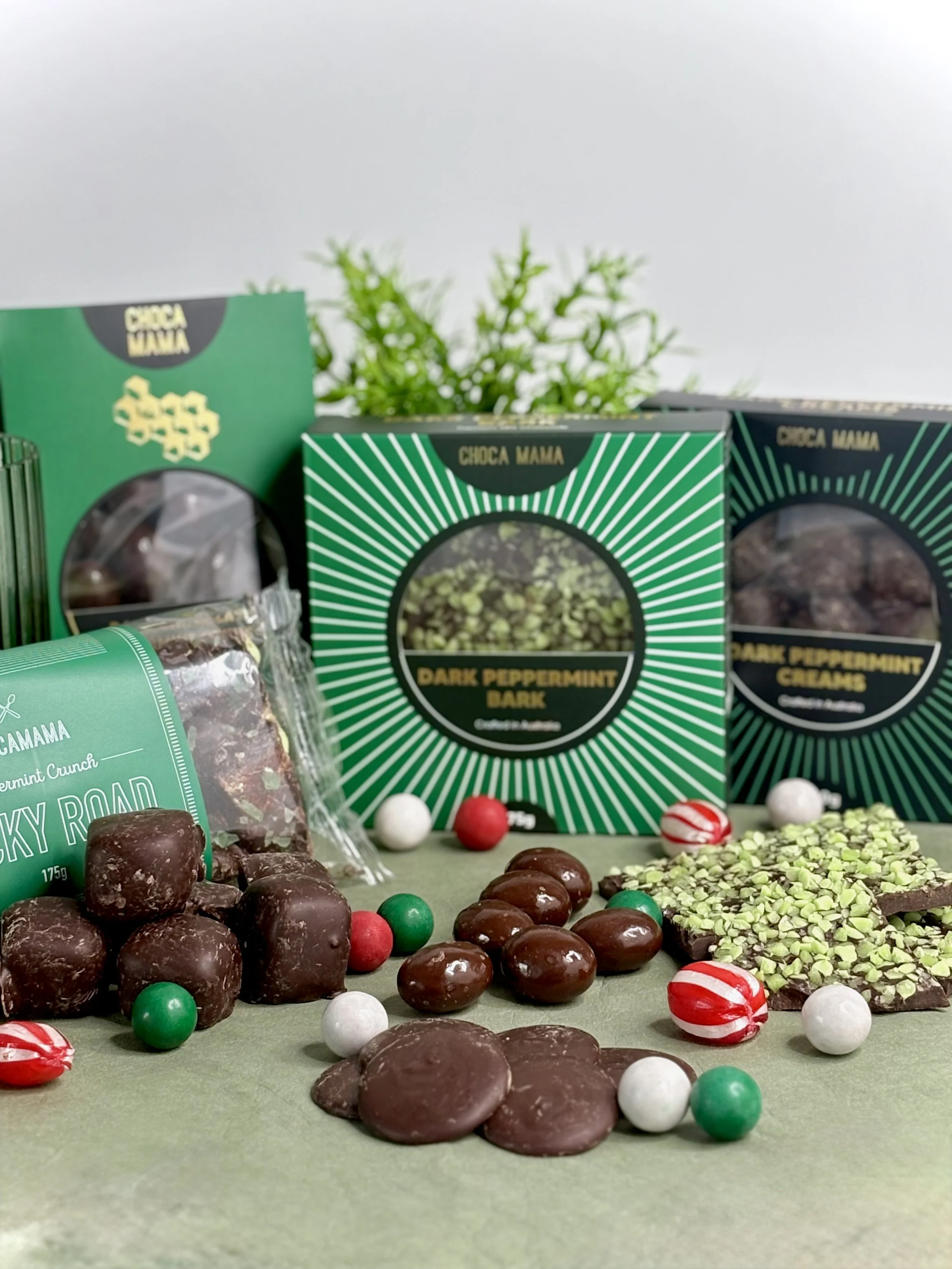 Assorted chocolates and candies on a green surface with decorative packaging and a plant background.