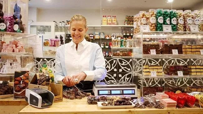 A woman smiling at a bakery counter with various chocolates and candies, including wrapped chocolates and gift boxes, in a well-lit shop.