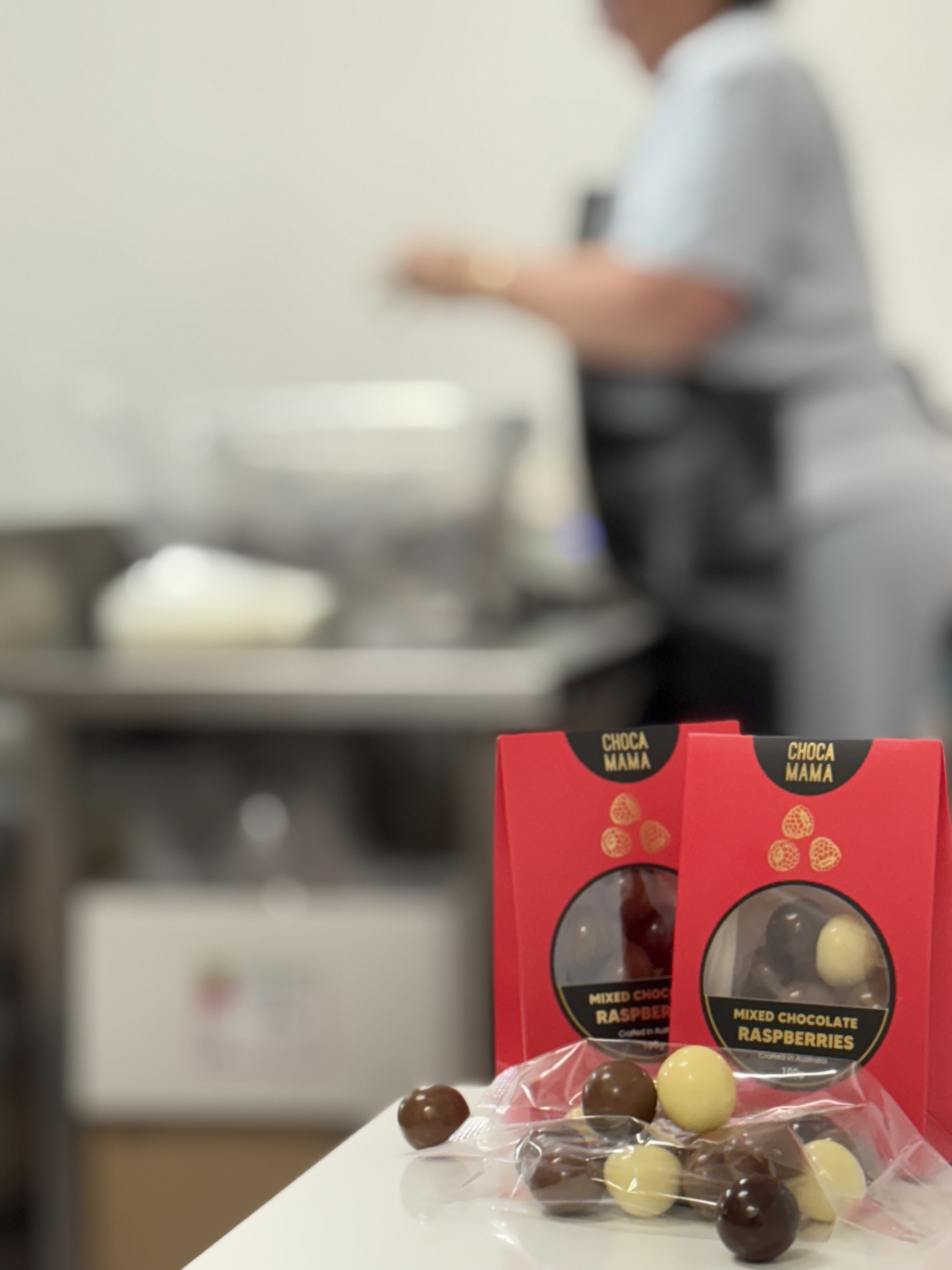 Close-up of a red box labeled "Choca Mama" with chocolate raspberry candies in front of it, out of focus background shows a person using a phone.