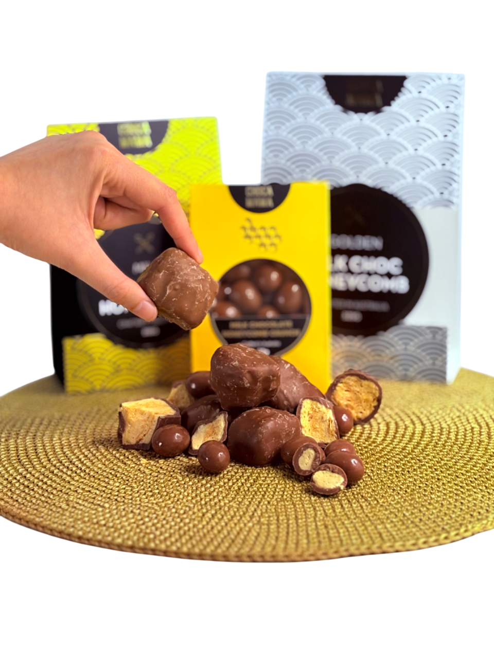 Hand holding a chocolate-covered caramel candy over a pile of assorted chocolates on a textured gold surface, with in the background two boxes of chocolates with black and yellow, and white and gray packaging.
