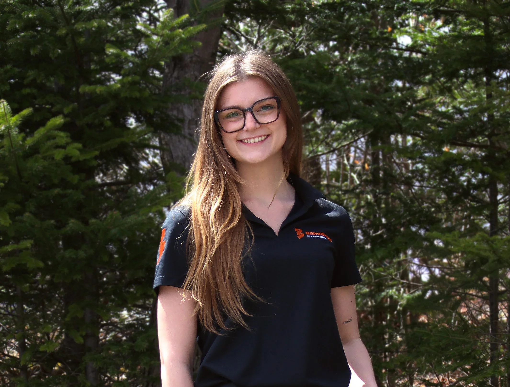 A young woman with long brown hair, glasses, and a black shirt standing outdoors in front of green trees, smiling at the camera.