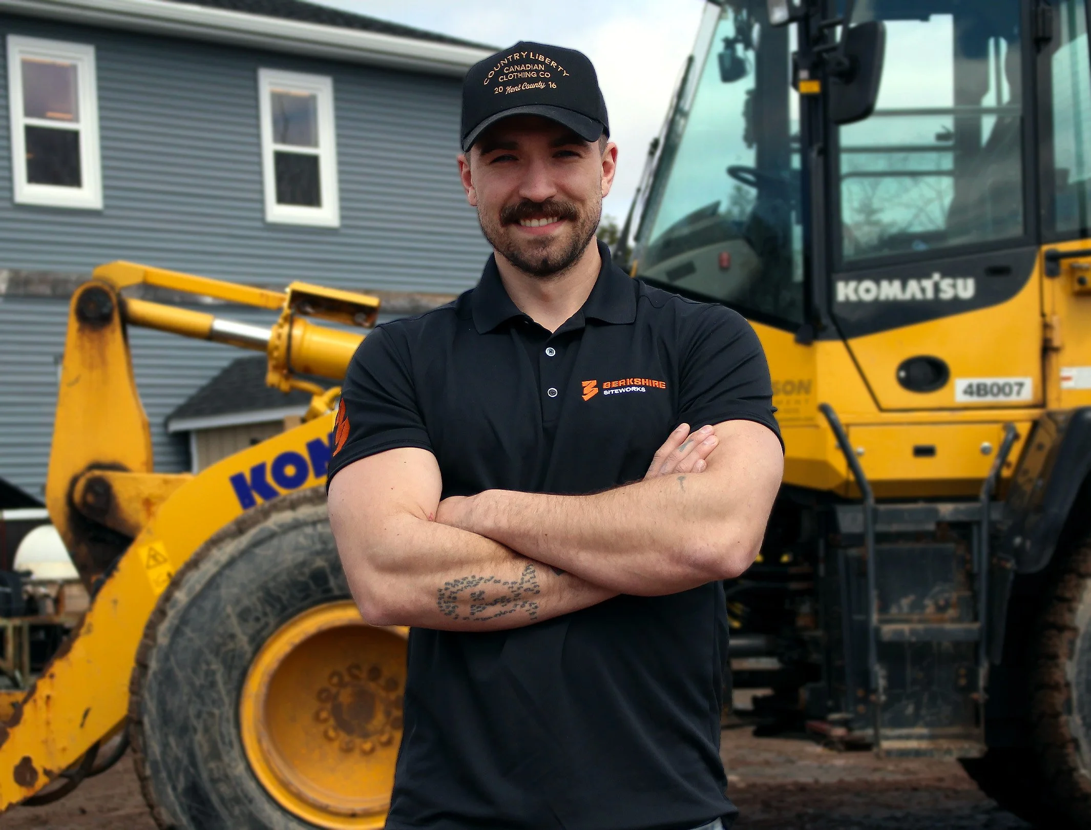 Man standing with arms crossed in front of yellow Komatsu construction vehicle, wearing a black polo shirt with logos, black cap, and sports tattoos on his forearm.