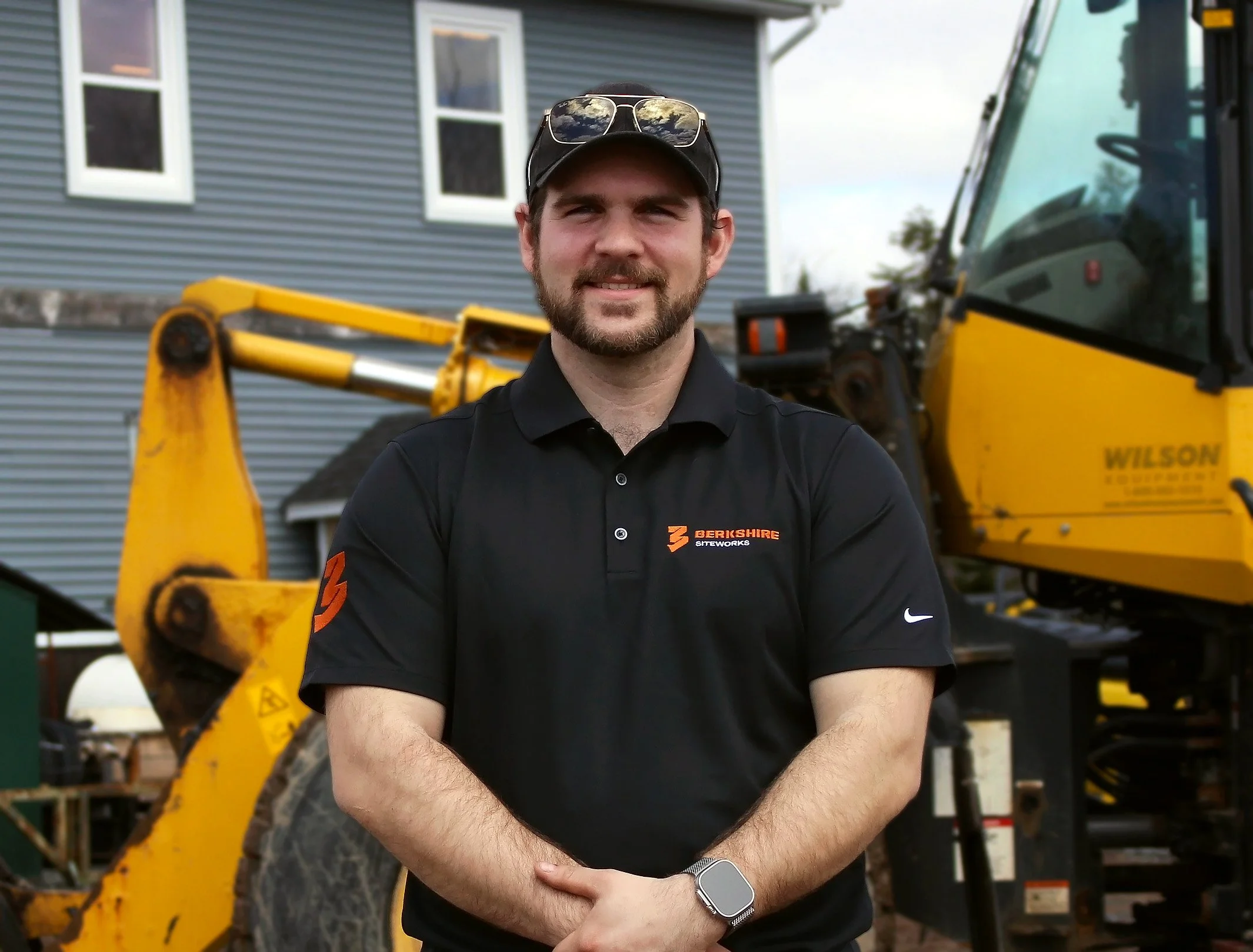 A man with a beard and sunglasses on his cap, wearing a black polo shirt with something written on it, standing in front of a yellow construction vehicle, posing with his arms crossed and smiling.