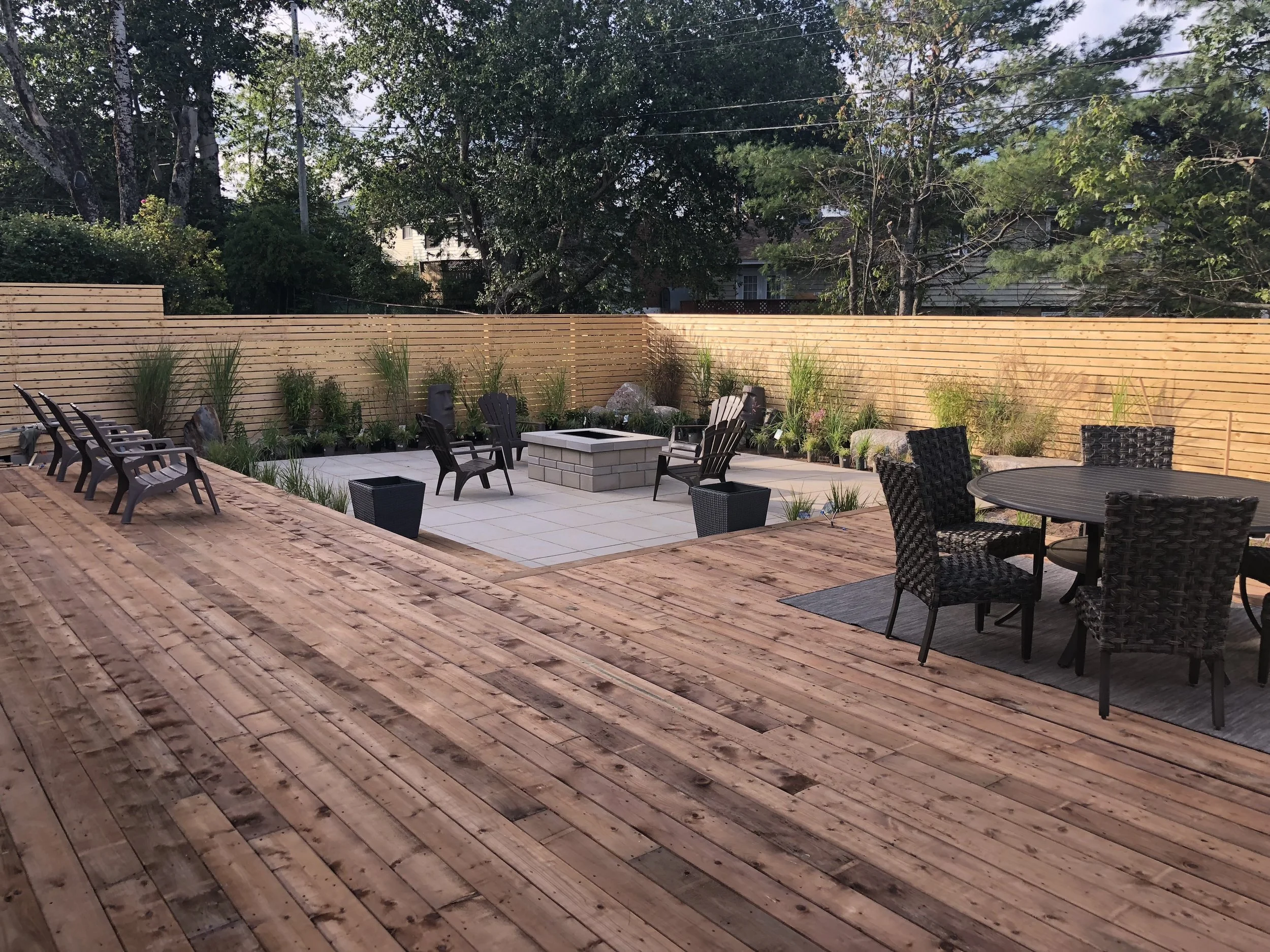 Backyard patio featuring a wooden deck, a fire pit area with Adirondack chairs, a separate seating area with wicker chairs and a round table, and a wooden privacy fence with plants and rocks along the perimeter.