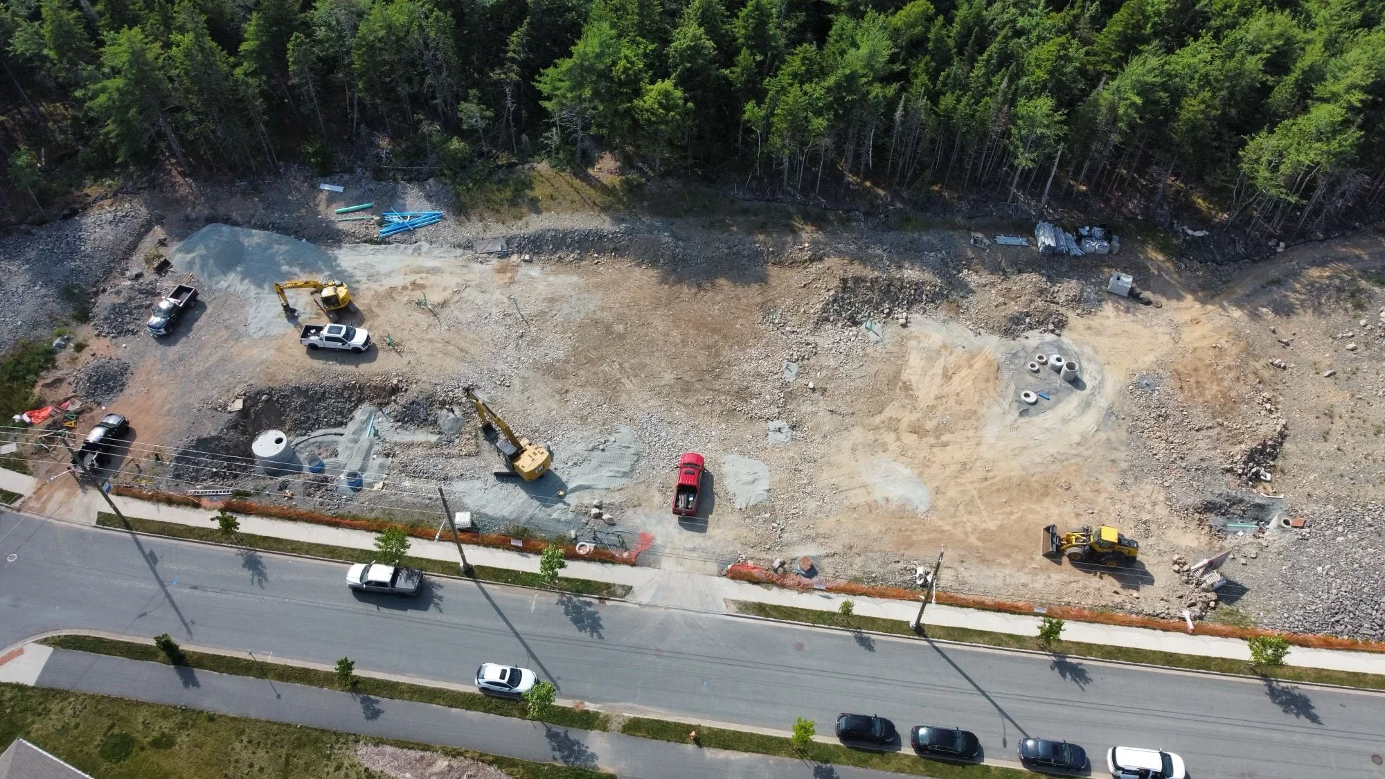 Aerial view of a construction site on a cleared plot of land with construction vehicles, pipes, and concrete cylinders, with a road and parked cars at the front and trees at the back.