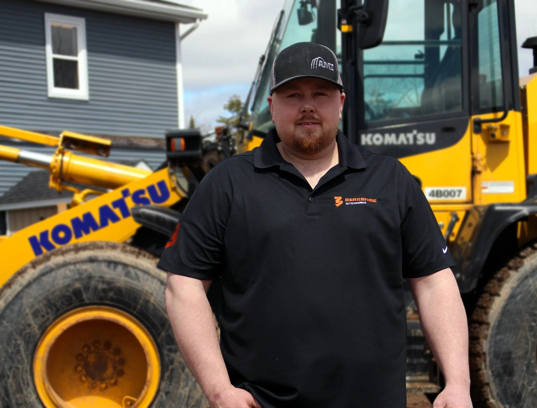 A man with a beard wearing a black polo shirt and baseball cap standing in front of a yellow Komatsu construction vehicle.