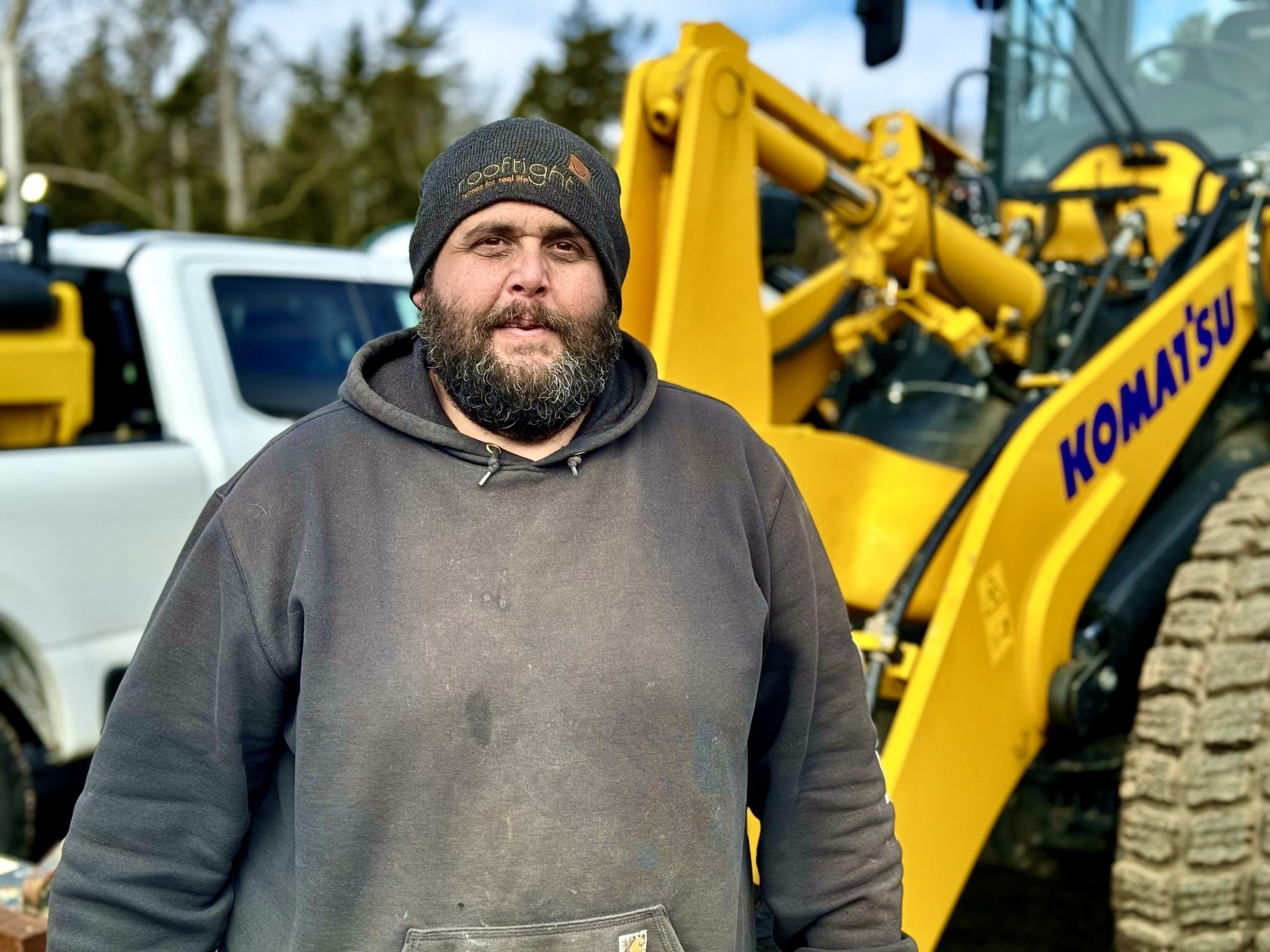 A man with a beard wearing a black beanie and a dark hoodie standing outdoors in front of yellow construction equipment. There are vehicles in the background.