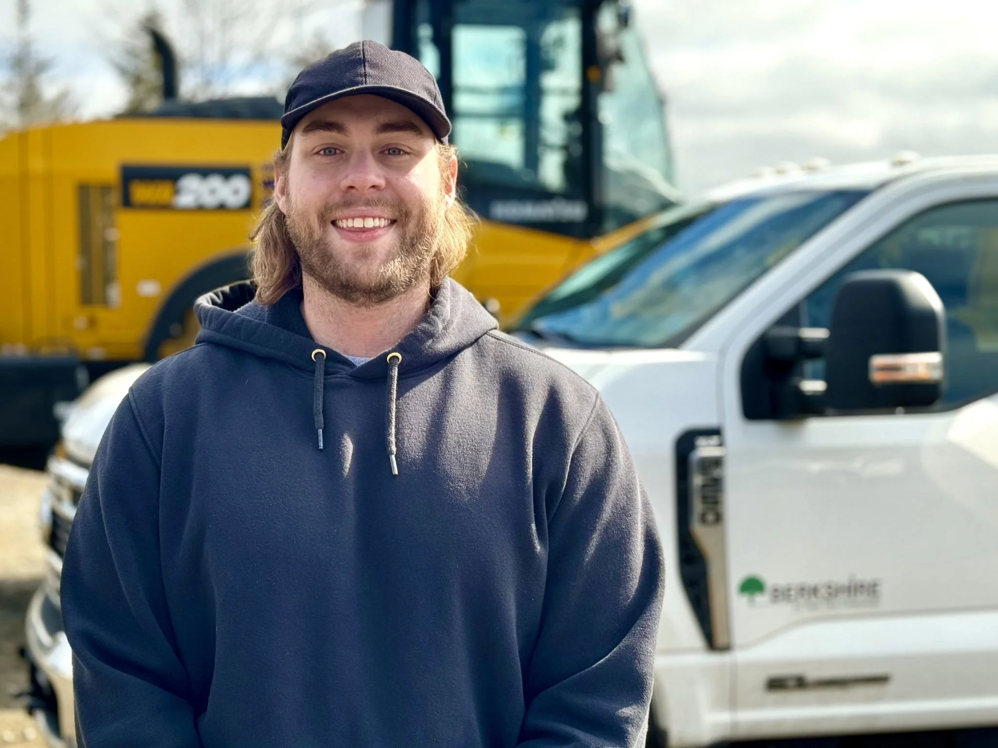 A man with long hair, a beard, wearing a dark hoodie and cap, smiling in front of construction vehicles and a white work truck.