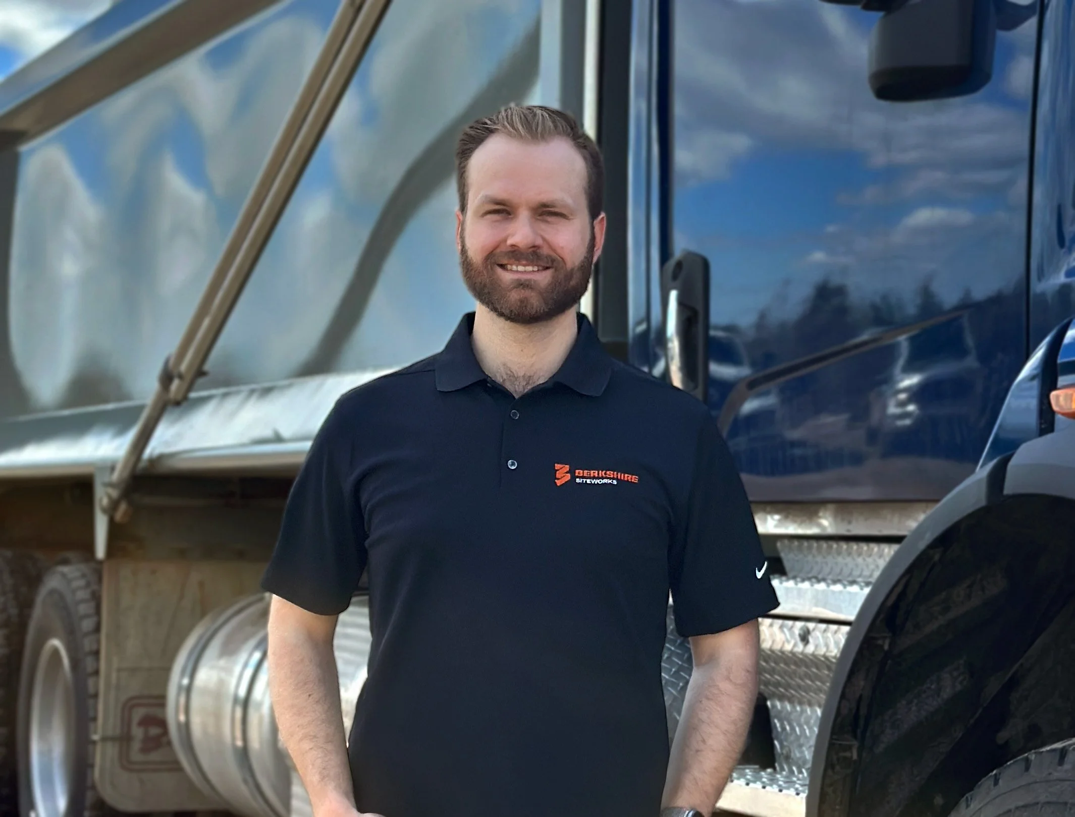 A man with a beard and short hair smiling, wearing a black polo shirt with a company logo, standing outdoors next to a large blue truck with shiny metal parts and a cloudy sky reflection.