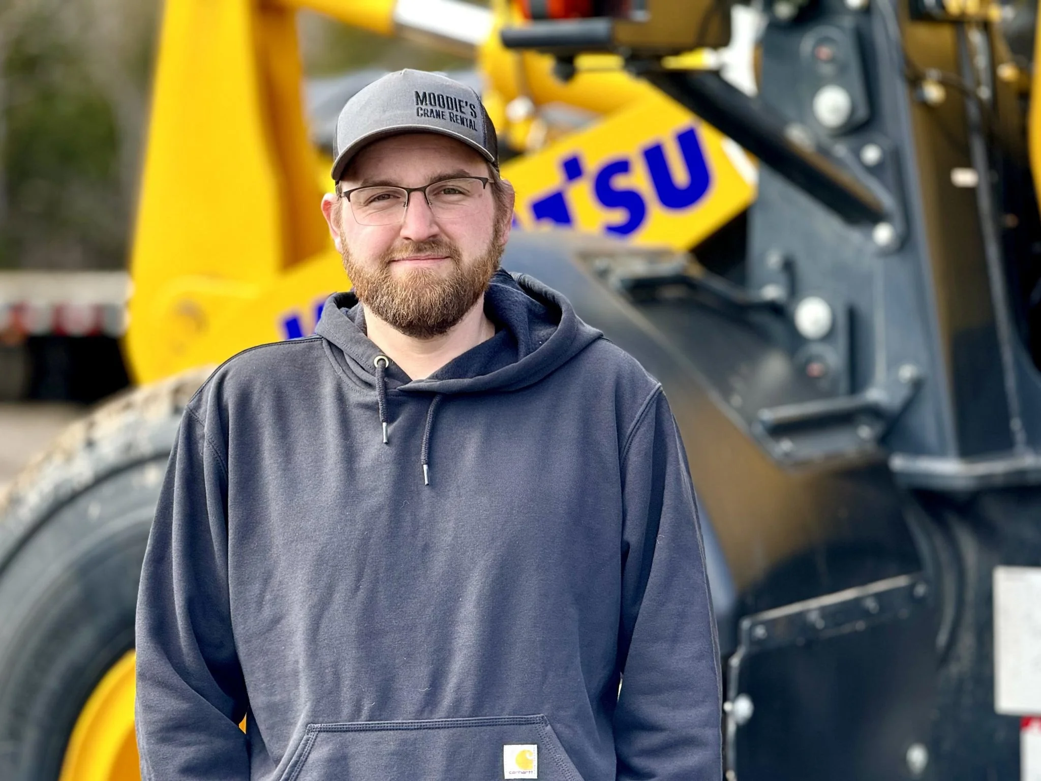 Young man with glasses and beard wearing a gray hoodie and cap, standing outdoors in front of yellow construction equipment with the word "UisU" visible.